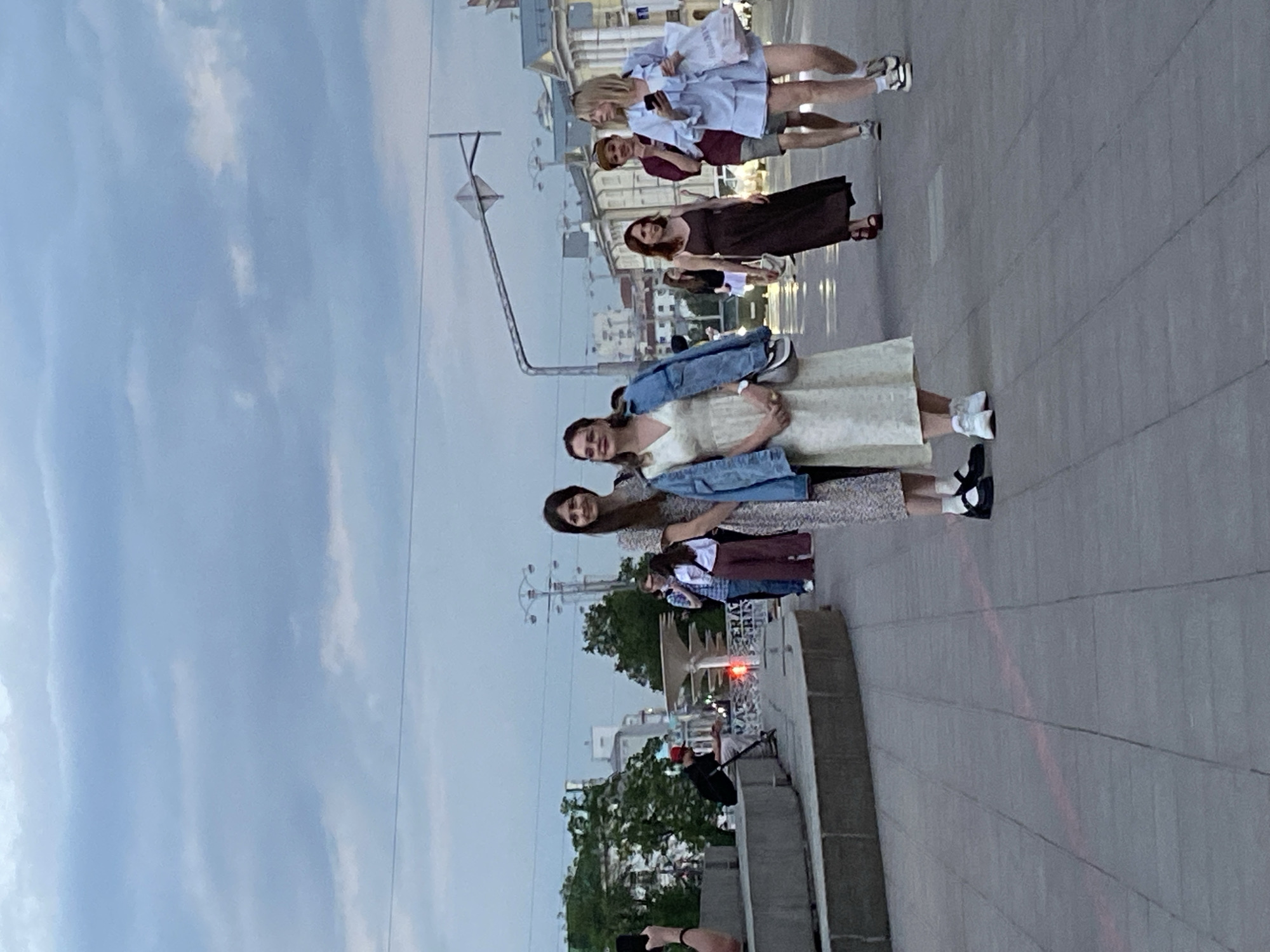 Several people are standing on a sidewalk, appearing to pose for a photograph. The image seems to be taken in an urban area, likely Yekaterinburg, Russia, as suggested by the architectural style and street fixtures. The background includes buildings, trees, street lights, and what looks like a Ferris wheel. The sky is overcast. A photographer is present at the bottom of the frame, holding up a phone to take a picture.