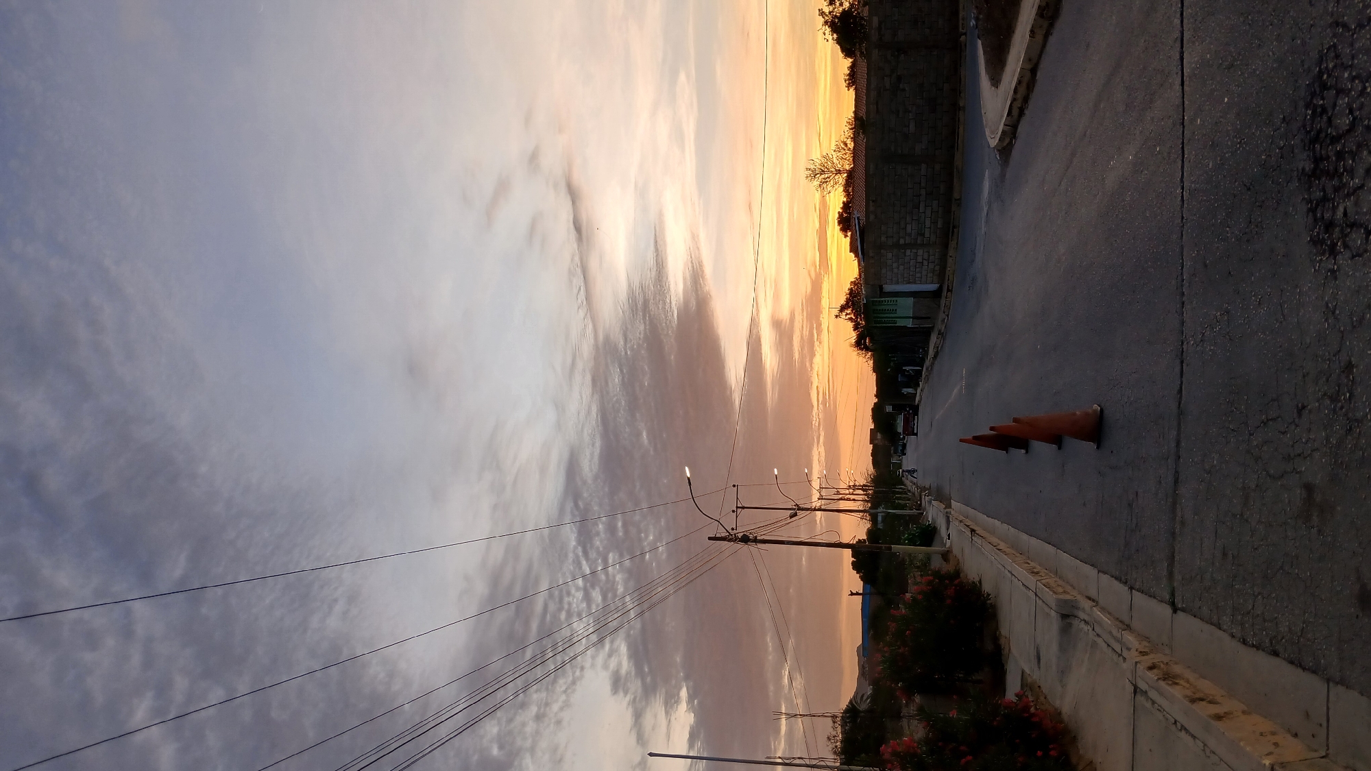 The image depicts a street in Altagracia, Venezuela at sunset. The sky transitions from shades of gray to orange. Power lines stretch across the sky, connected to streetlights that line the street. On the right, a paved road is bordered by a concrete curb. Three orange traffic cones are situated on the road. A low wall separates the road from what appears to be residential properties. Trees and foliage are visible behind the wall. The overall scene suggests a quiet residential area during the evening hours.