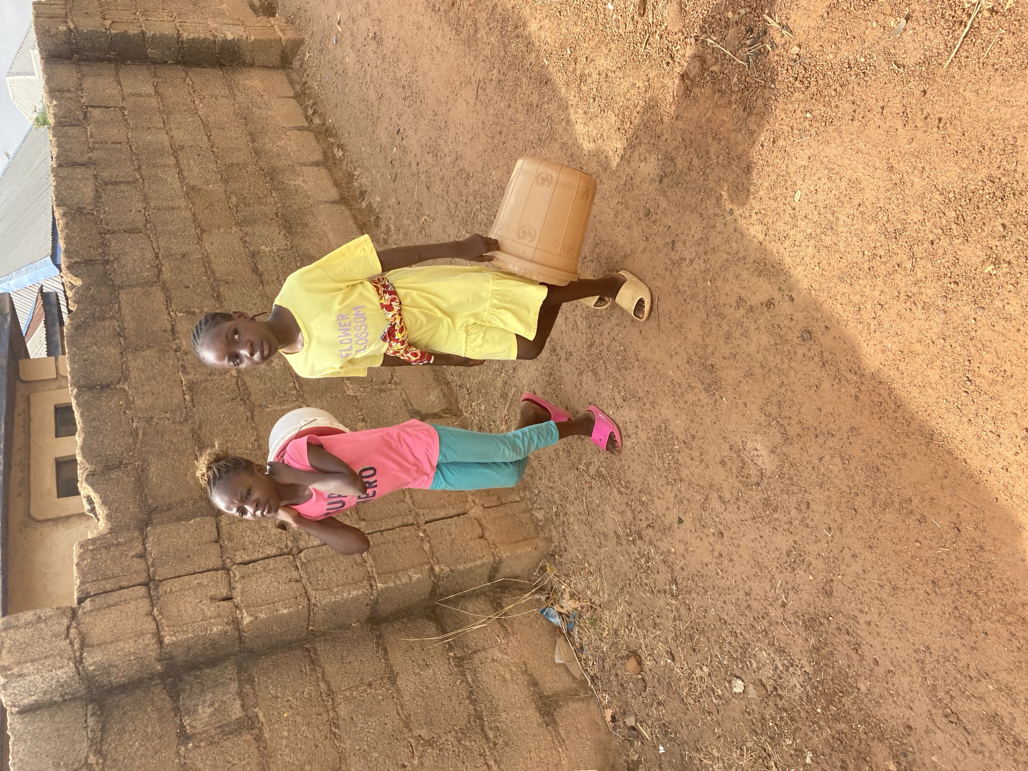 Two young girls stand on a dirt path next to a block wall in what appears to be an arid location. One girl wears a yellow dress, sandals, and holds a plastic bucket. The other girl wears a pink shirt, blue leggings, pink sandals, and has a white bucket on her head. The location is suggested to be Bukuru, Nigeria. The setting appears to be a residential area with visible buildings. The ground is dry, with dirt and scattered debris. The girls are looking towards the camera.