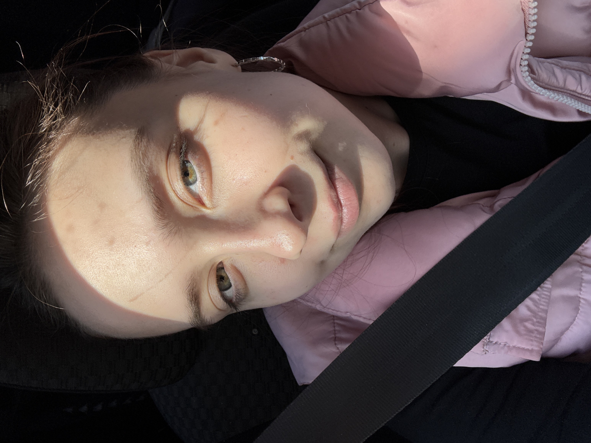 A young woman is depicted in a close-up portrait, reclining or lying down, with her head tilted to her right. She has fair skin, light brown hair, and green or hazel eyes, which are looking directly at the camera. A gentle, subtle smile is present on her lips. Strong, direct sunlight illuminates parts of her face, creating a striking pattern of light and shadow across her forehead, nose, and cheeks. Some fine strands of hair are visibly catching the light.
She is wearing a light pink quilted jacket with a white zipper, layered over a dark or black top. A prominent black seatbelt diagonally crosses her upper body, indicating she is likely inside a vehicle, such as a car. A silver hoop earring is visible in her left ear. The background of the image is dark and out of focus, keeping the viewer's attention on the woman. The strong sunlight suggests it is daytime with clear, sunny weather. There is no visible text in the image. The setting is Votkinsk, Russia.