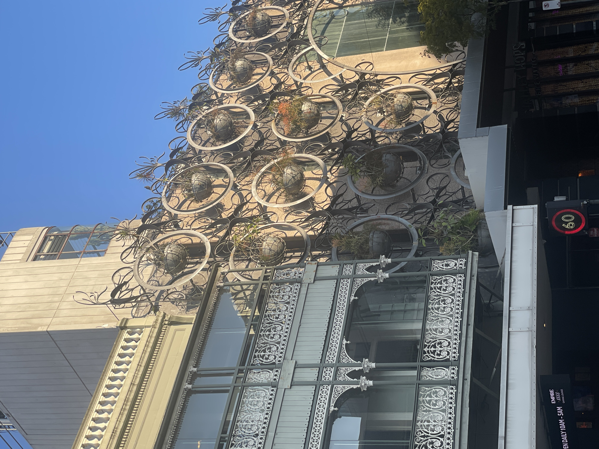 The image shows the exterior of a building located in Fortitude Valley, Australia, with a clear blue sky overhead. The building features a distinctive facade adorned with a decorative metal installation resembling a series of interwoven rings, some of which hold hanging planters with greenery. To the left, there is a section of the building with a detailed white cast-iron balcony. Below this balcony, a sign indicates "OPEN DAILY 10AM - 5AM EMPIRE 1888 GAMING LOUNGE". In the foreground, a traffic light displaying the number "60" in red is visible, along with a street sign. The overall scene captures an urban architectural style with ornamental elements and clear daytime lighting.