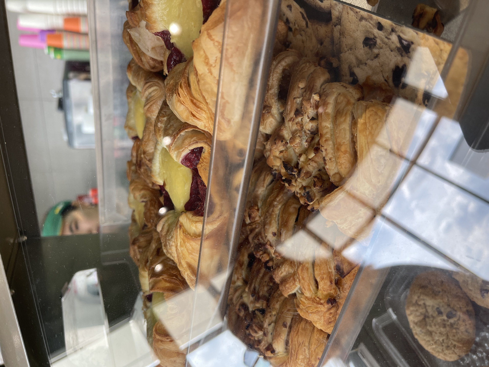 A bakery display case containing pastries is shown. On the left are pastries filled with red jam and yellow custard. Next to them are pastries with nuts, and on the right are chocolate chip cookies. A woman with a green head covering is reflected in the glass. The location is unspecified but can be assumed to be Russia, Moscow.