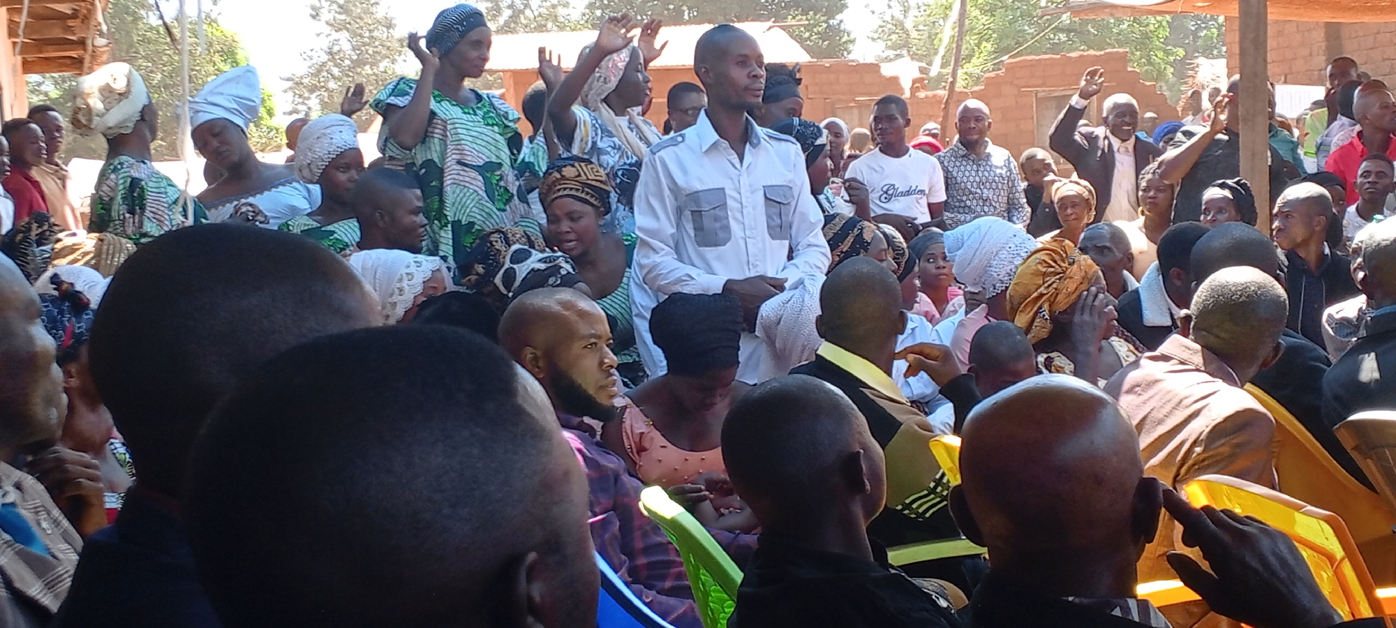 The image depicts a gathering of people in what appears to be an outdoor setting, possibly in Kalemie, Democratic Republic of the Congo. The majority of the people are seated, while others stand in the background. The crowd seems to be diverse in age and attire. Many of the women are wearing traditional head coverings, and their clothing is a mix of colorful, patterned fabrics and more contemporary styles. A man in a light-colored shirt stands out in the center of the crowd. Some people are raising their hands, indicating a possible gesture of participation or greeting. The environment consists of trees and simple structures.