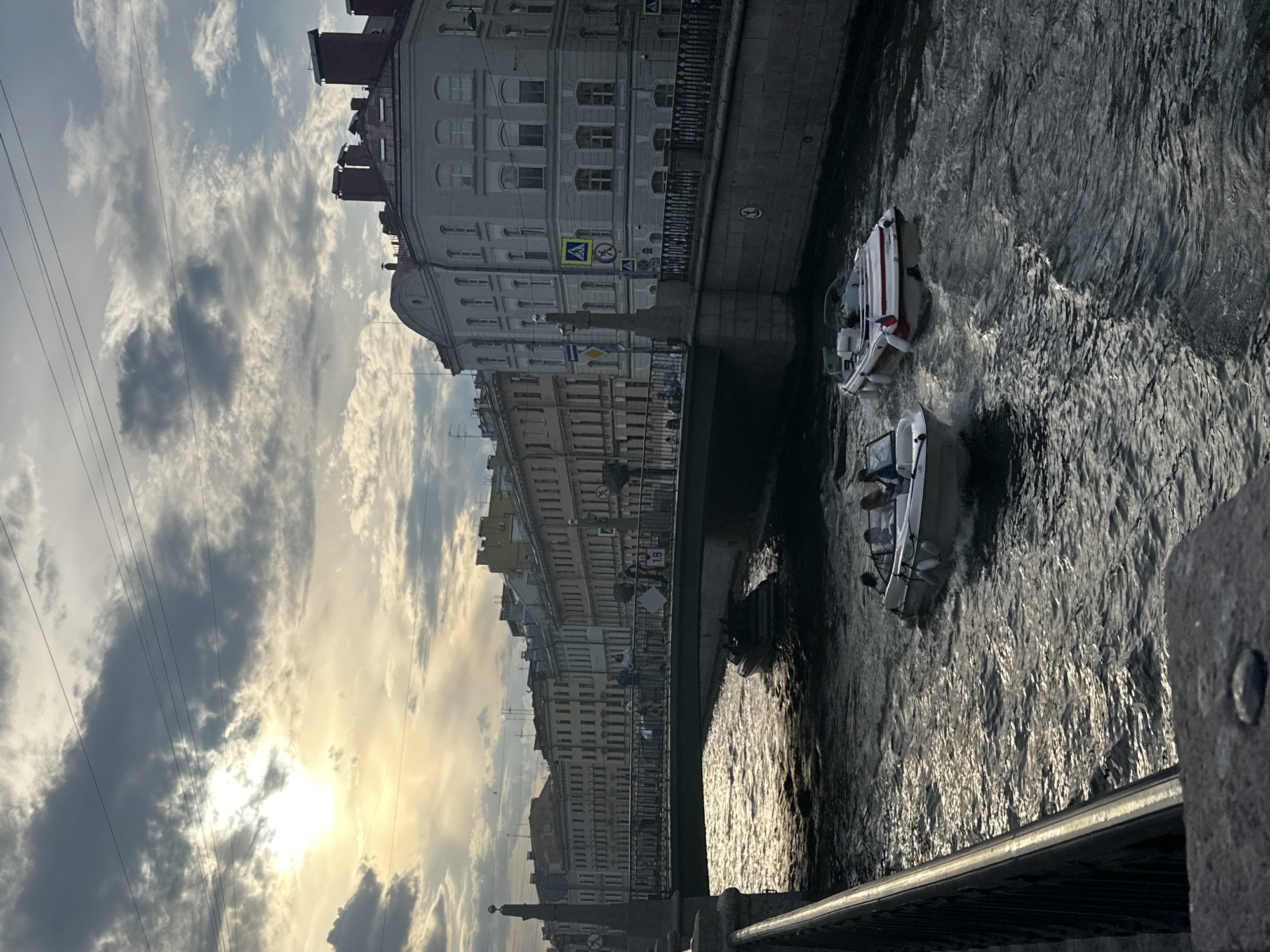 The image depicts a canal scene in Saint Petersburg, Russia, during golden hour, with the sun low and partially obscured by clouds in the upper left quadrant. The central focus is a waterway with two motorboats in the foreground, approaching the viewer.
The sky exhibits a mix of bright, sunlit clouds and darker, heavier cloud formations. Several thin, dark electrical wires traverse the upper portion of the sky horizontally.
The canal water is dark and choppy, reflecting the sun's light in shimmering patterns, particularly towards the left side of the frame.
Two open-top motorboats are prominent. The boat on the left is light-colored and carries at least three individuals, two of whom are visible in the bow area. It is moving towards the viewer, creating a wake. The boat on the right is white with red and white markings along its hull and prominently displays what appears to be the national flag of Russia (white, blue, red horizontal stripes) at its stern. This boat is also moving, generating a distinct wake. A third, darker boat is partially visible further down the canal, beneath the bridge.
A multi-arched stone bridge with a dark green metal railing spans the canal. Several pedestrians are visible walking across the bridge; one person on the right side of the bridge is holding a white rectangular object with the number "18" visible. Stone balustrades with decorative finials mark the bridge's edges.
Both banks of the canal are lined with historic, multi-story buildings, typically four to five stories tall, featuring classical architectural styles, including arched windows on lower levels, cornices, and decorative facades. Building colors vary from light gray to beige and light yellow. Reddish-brown roofs with multiple prominent chimneys are visible above the building lines. On the right, a distinctive light-gray corner building with a rounded facade faces the canal. Various road signs, including a yellow pedestrian crossing sign, are visible on poles and buildings.
In the immediate foreground, a dark, textured stone balustrade or railing of the bridge from which the photograph was taken extends diagonally from the lower left towards the center. A single metal stud is visible on the top surface of this stone structure in the lower left.