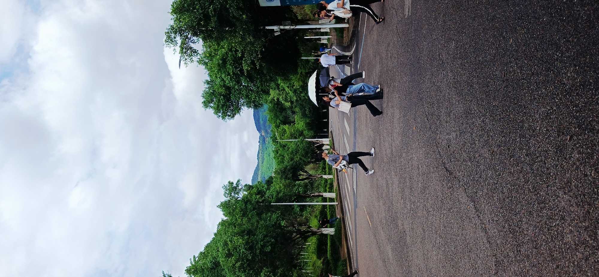 The photograph shows a street scene in Chongqing High-tech Zone, China. Several people are crossing a paved road, some with an umbrella. Trees line the side of the road, and mountains are visible in the background. The sky is overcast.