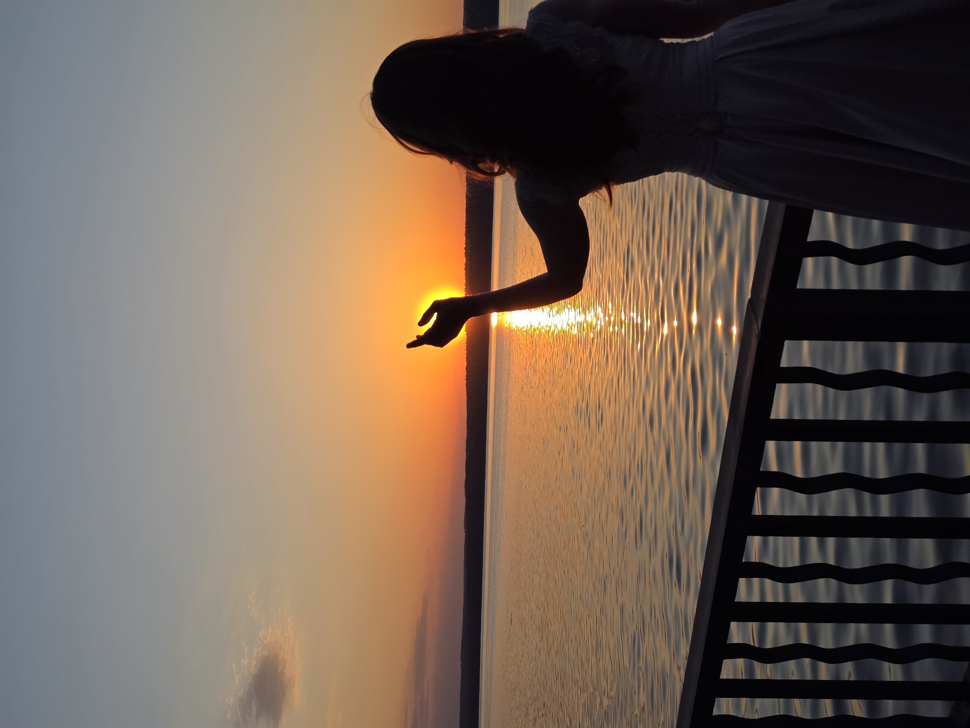 The image depicts a woman silhouetted against a sunset over a body of water. She is leaning over a railing, and her hand is positioned as if she is touching the sun. The railing appears to be part of a pier or boardwalk. The sky graduates from a light blue to a deep orange and red at the horizon, where the sun is setting. The water reflects the colors of the sky. Given the prompt about Chaikovsky, Russia, it is highly probable that this image was taken at the Kama Reservoir, given its prominence in the area.
