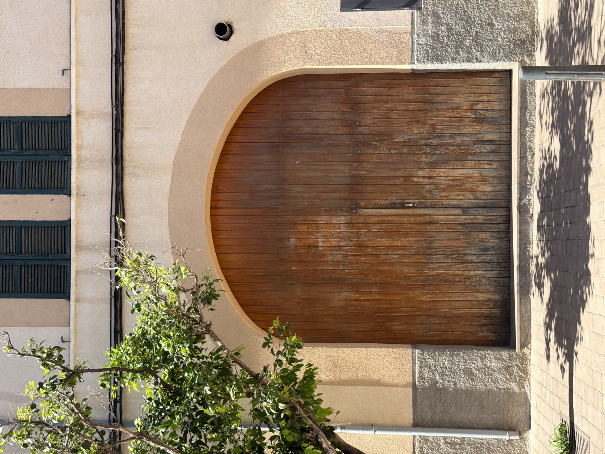 A large, weathered, wooden arched double door is centered in the image. The door is made of vertical wooden planks and has two handles. Above the door, a light peach-colored wall features two dark green shutters on a window. Black electrical cables run horizontally across the wall above the door, and a small round ventilation pipe is visible to the right of the arch. To the left of the door, a young tree with green leaves and small green fruits is partially visible, leaning towards the building. A gray downspout runs vertically along the building's wall to the left of the tree. The lower portion of the building wall, beneath the arch, is a rough, grey textured surface. The ground in front of the building is a light brown brick-paved sidewalk, with shadows from the tree and overhead foliage cast upon it. A small patch of green grass and a drain grate are visible on the lower left of the sidewalk. A dark gray bollard stands on the sidewalk to the right of the frame. The scene appears to be an exterior facade of a building in a Mediterranean setting, likely in Manacor, Spain, given the architectural style and vegetation.