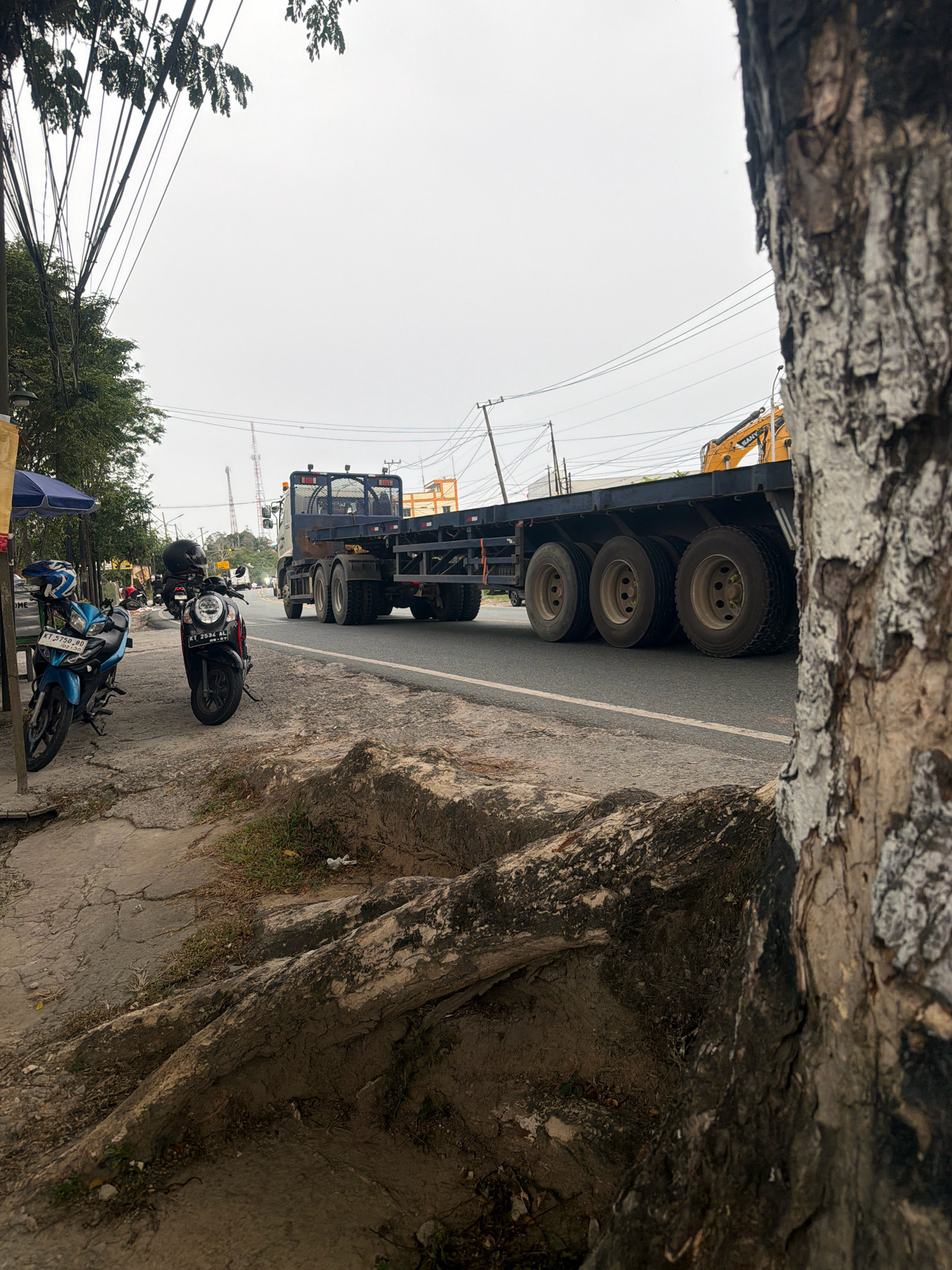 The image depicts a street scene in Balikpapan, Indonesia, with a focus on a large flatbed truck carrying a yellow excavator. The truck is in motion on a paved road, with its rear wheels prominently visible. To the left of the road, there is a sidewalk with two parked motorcycles, one blue and one black, and a small blue umbrella covering a stand. Trees and utility poles with numerous wires line the street, indicating an urban or semi-urban environment. The foreground features exposed tree roots and cracked pavement, suggesting an older or less maintained area. The sky is overcast, contributing to the muted lighting of the scene. The overall impression is a snapshot of daily life and infrastructure in a Southeast Asian city.