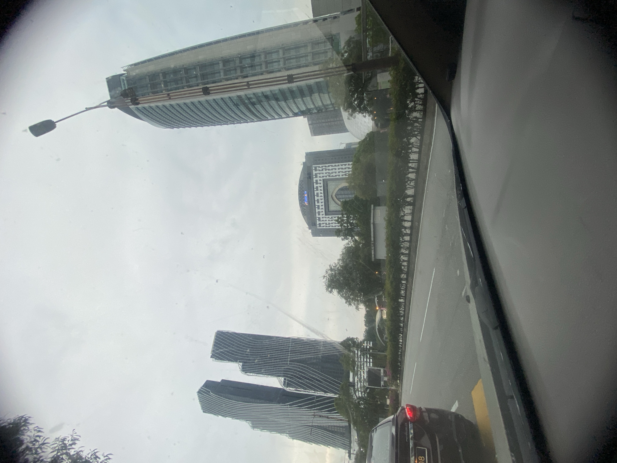 This image captures a view from inside a vehicle on a cloudy day in Kuala Lumpur, Malaysia. The foreground shows the car's dashboard and windshield, which has some smudges or water droplets. In the midground, a dark-colored car is visible in the left lane of a multi-lane road. The road is lined with a dense hedge.
The background is dominated by several prominent modern skyscrapers. To the left are two strikingly designed, wavy skyscrapers with a dark facade and white vertical lines. Further to the right, a tall, slender skyscraper with a green-tinted glass exterior stands out. In between, there is a building with a distinctive arched facade that resembles a traditional Islamic architectural element, featuring geometric patterns and the word "MAXIS" visible at the top. Another building with a curved, dome-like structure is also visible. The sky is overcast, suggesting potentially poor weather conditions. The overall impression is an urban landscape characterized by contemporary architecture.