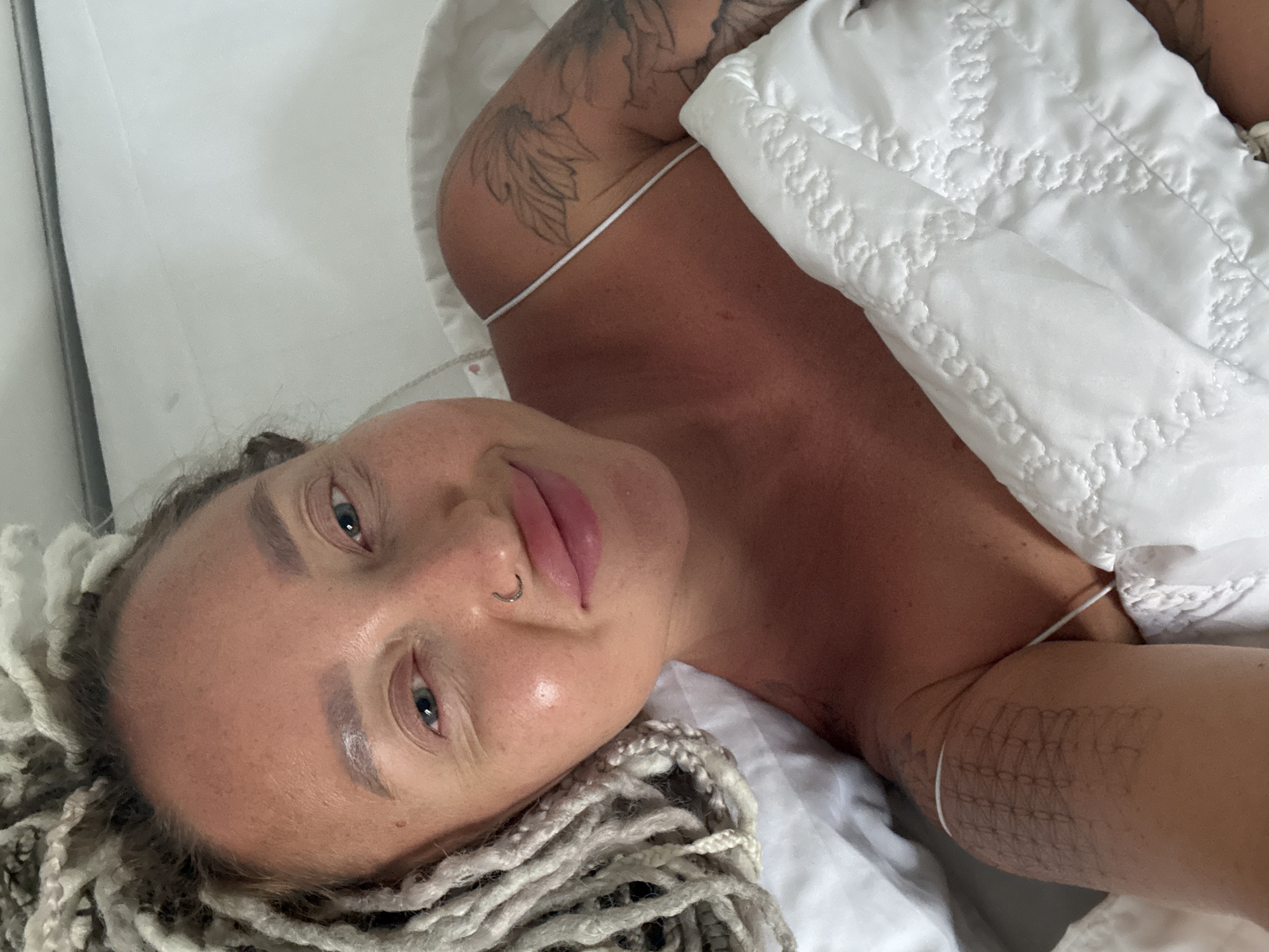 A close-up, high-angle shot shows a woman lying in bed, covered by a white quilted duvet. She has dreadlocks, a nose ring, and tattoos on her arms and shoulder. Her skin appears tanned. The background is white, suggesting a bedroom setting. The image does not contain any information that can identify its specific location within Bac Nha Trang Ward, Vietnam.