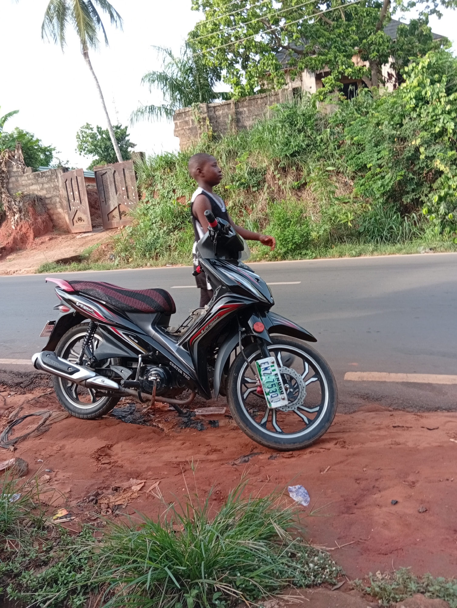 In Nkpor, Nigeria, a young person stands near a parked, black and red Carter Tec motorcycle on the side of a paved road. The motorcycle has a visible license plate, "ENZ-7530J," and is partially situated on a patch of reddish-brown soil with sparse vegetation and litter. In the background, lush green vegetation covers a small hill, with a building partially visible behind the foliage. A palm tree and a gated brick wall stand to the left of the scene. The individual is dressed in dark clothing with a light-colored tank top and appears to be looking to the right of the frame.