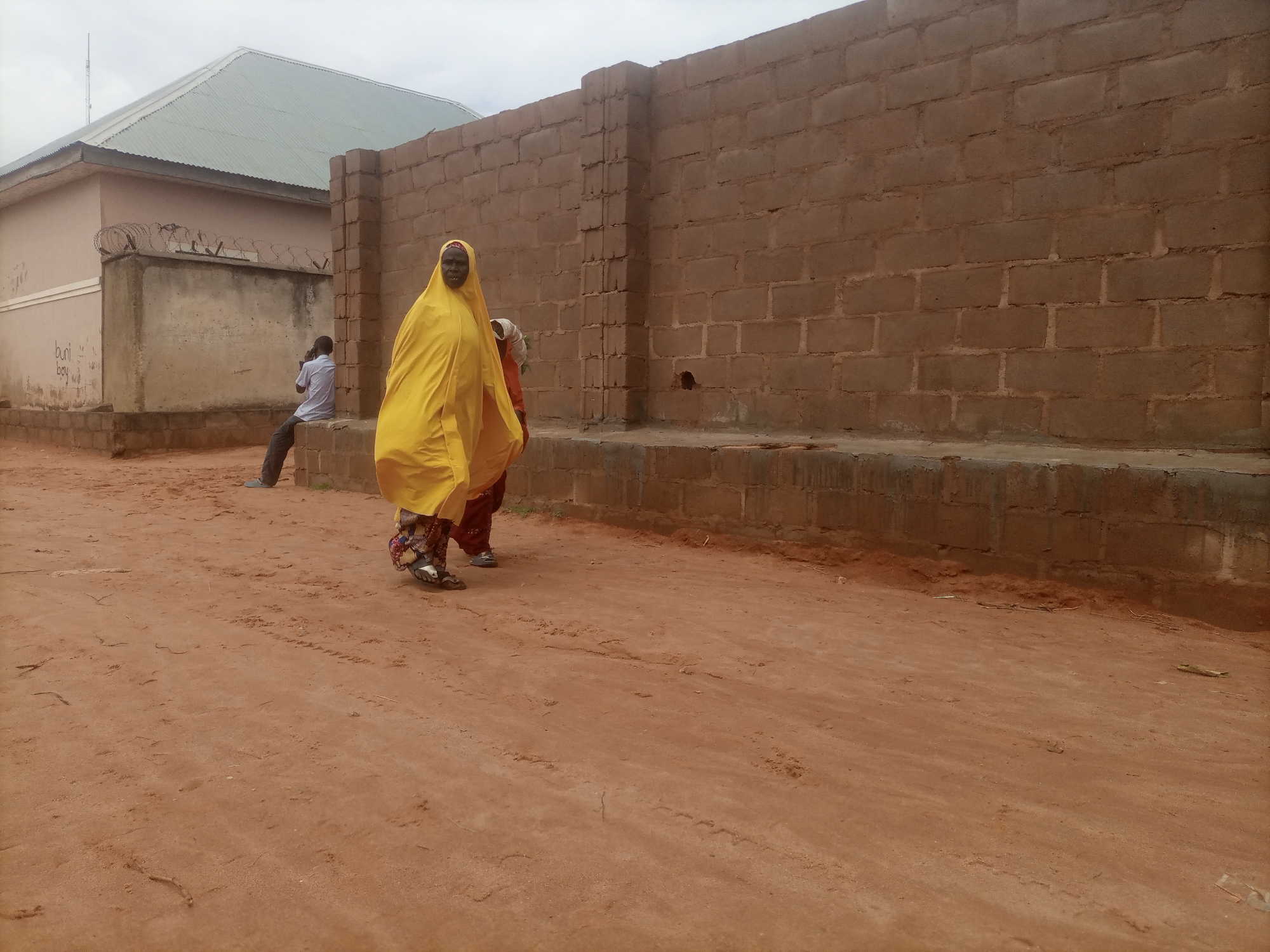 In Tumfure, Nigeria, a two-story building with a metal roof is visible to the left, bordered by a low concrete wall topped with barbed wire. A man in a light blue shirt and dark pants sits on the wall. In the foreground, two women are walking on a dirt path. The woman on the left wears a bright yellow head covering and a patterned skirt. The woman on the right wears an orange head covering and a dark skirt. A large brick wall runs along the right side of the image. The ground is sandy and reddish-brown, with some scattered debris.