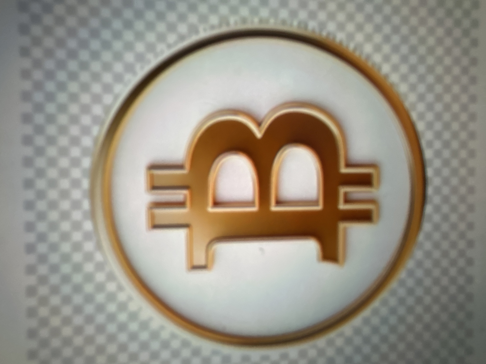 The image shows a close-up of a golden Bitcoin symbol on a white background with a subtle gold outline. The background behind the Bitcoin symbol is a blurred, checkered pattern of light gray and white squares, suggesting a transparent or digital background. The Bitcoin symbol itself is a stylized capital letter 'B' with two vertical lines running through its center, rendered in a metallic gold color with a subtle gradient and shine, giving it a three-dimensional appearance. The image is slightly out of focus. There is no discernible information about the city of Gelendzhik, Russia, or any people, actions, or specific environmental details related to a physical location.