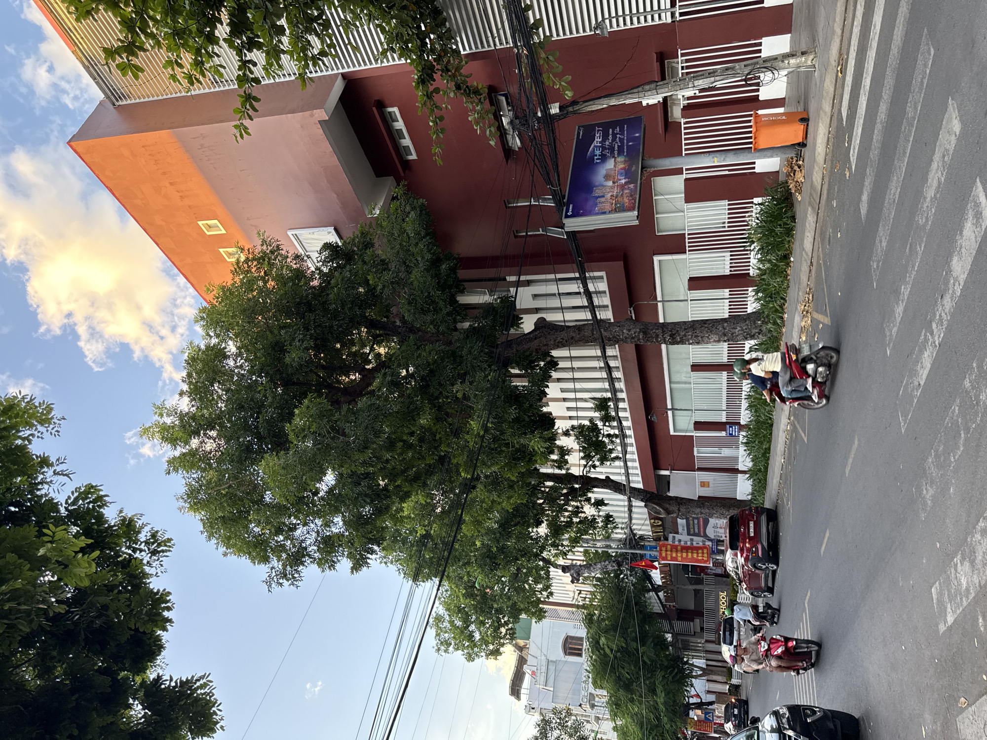 In Nha Trang, Vietnam, a street scene unfolds under a partly cloudy sky, with a prominent red building dominating the right side of the frame. Large trees with dense green foliage are interspersed with the urban architecture, casting shadows on the road and sidewalk. Several motorcycles navigate the asphalt street, some with multiple passengers. A red car is parked on the side of the road. Across the street, a white building with a "SCHOOL" sign is visible. A billboard advertising "THE FEST" stands near the red building, accompanied by an orange trash bin on the sidewalk. Power lines crisscross the sky, adding to the urban character of the scene.