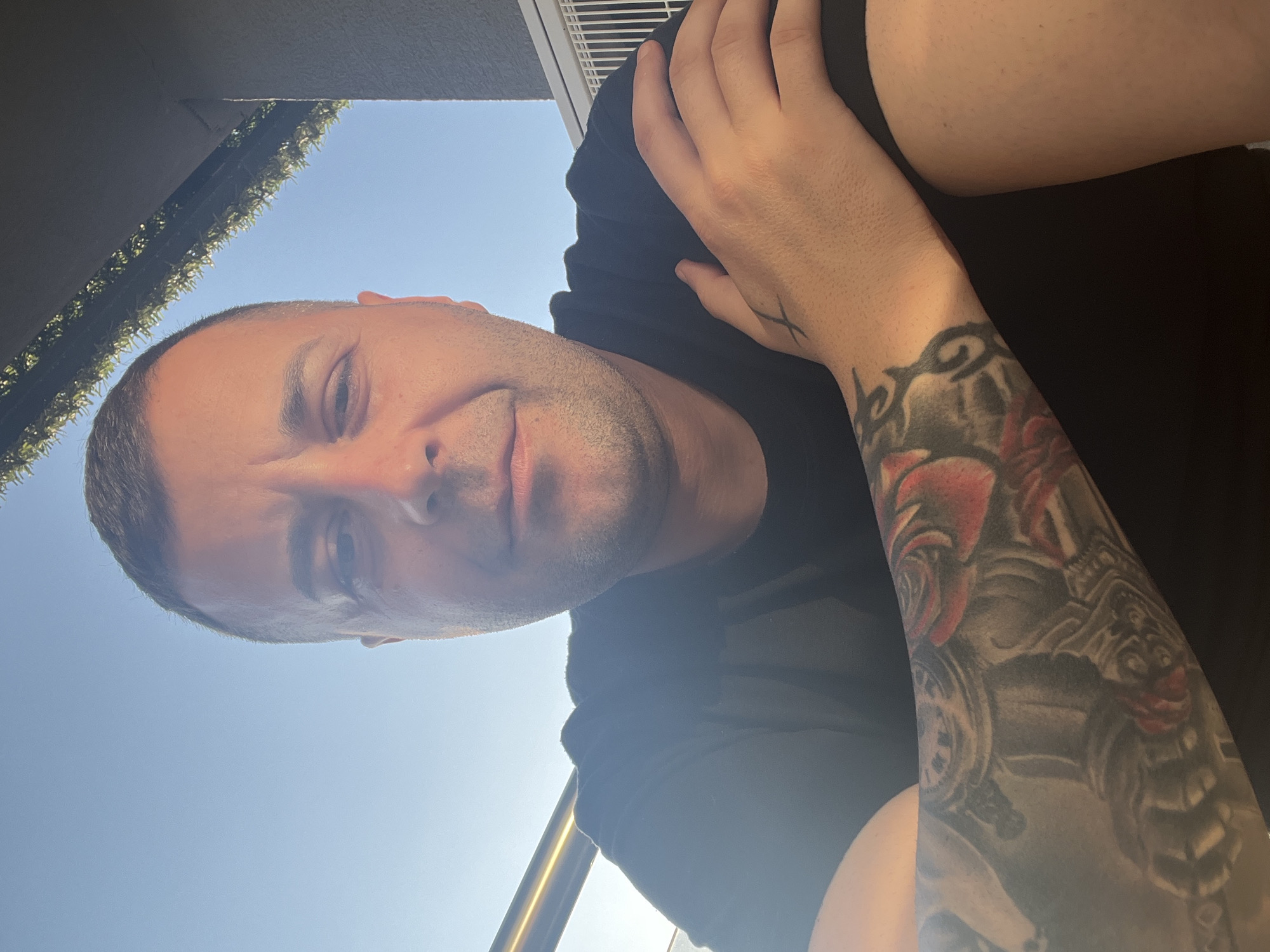 A man is pictured from a low angle against a backdrop of a clear blue sky. A glimpse of greenery and a white railing can be seen above and to the left of his head. He has short hair and stubble. He is wearing a black t-shirt. His left arm is positioned across his chest and has intricate tattoos on the lower arm and a small tattoo on the hand. The location is speculated to be Varna, Bulgaria, although there is no definitive visual information to confirm this beyond the context of the request.