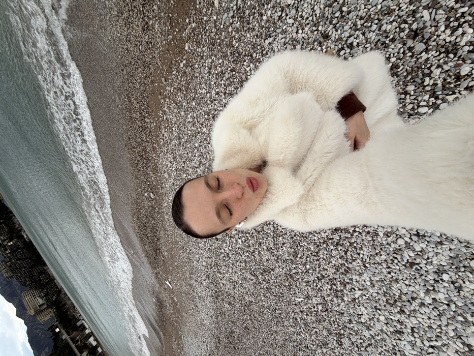 An overhead shot captures a person lying on their back on a pebble beach, next to the sea. The image is tilted approximately 45 degrees, making the horizon line run diagonally from the bottom left to the upper right.
The central subject is a person, likely an adult, with their eyes closed, suggesting they are resting or asleep. Their facial features are relaxed, with naturally colored lips slightly parted. Their dark hair is pulled back, revealing a clean forehead and temples. They appear to have fair skin.
They are dressed in a prominent, voluminous, off-white or cream-colored fluffy coat or garment that covers their entire body, blending somewhat with the lighter pebbles of the beach. A dark, possibly brown or burgundy, long-sleeved garment is visible peeking out from under the cream coat on their right arm. A small, shiny detail, possibly an earring, is visible on their left earlobe.
The setting is a stony beach composed of various shades of grey and white pebbles, which are clearly visible beneath and around the person. To the left of the person, gentle waves break on the shoreline of a calm, greenish-blue body of water, which appears to be the sea.
In the far background, on the left side of the image, there are buildings and what looks like hills or mountains, indicating a coastal landscape. The sky above appears overcast, providing soft, diffused lighting to the scene. The overall mood conveyed is one of peace and tranquility. There is no visible text in the image.