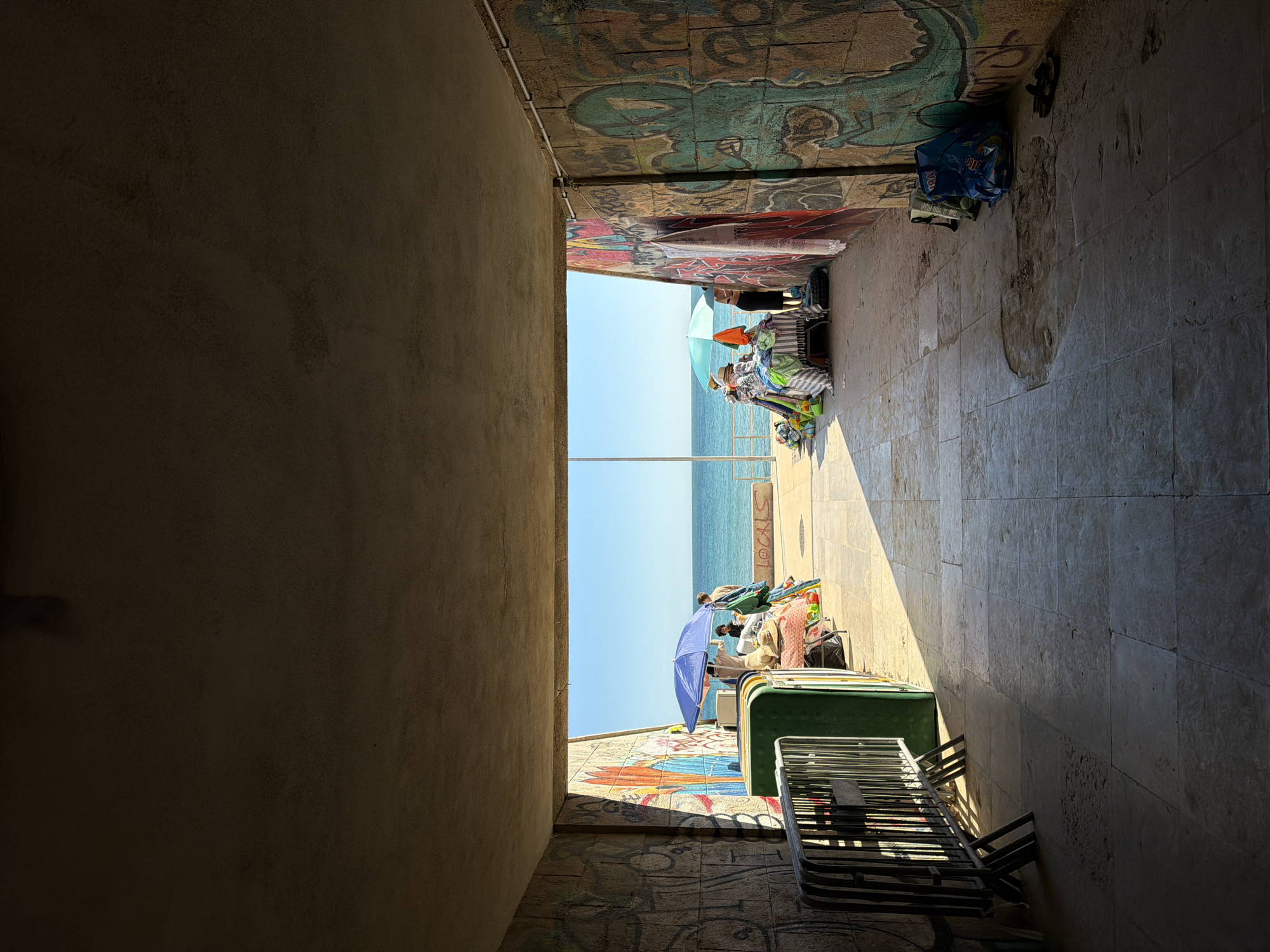 The image shows a view from a dark, narrow passage in Cascais, Portugal, towards a sunlit beach. The passage walls are adorned with graffiti. People are visible on the beach, sitting under umbrellas. Beach chairs and other beach equipment are visible within the passage. The sea is calm and blue under a clear sky. The ground of the passage is tiled.