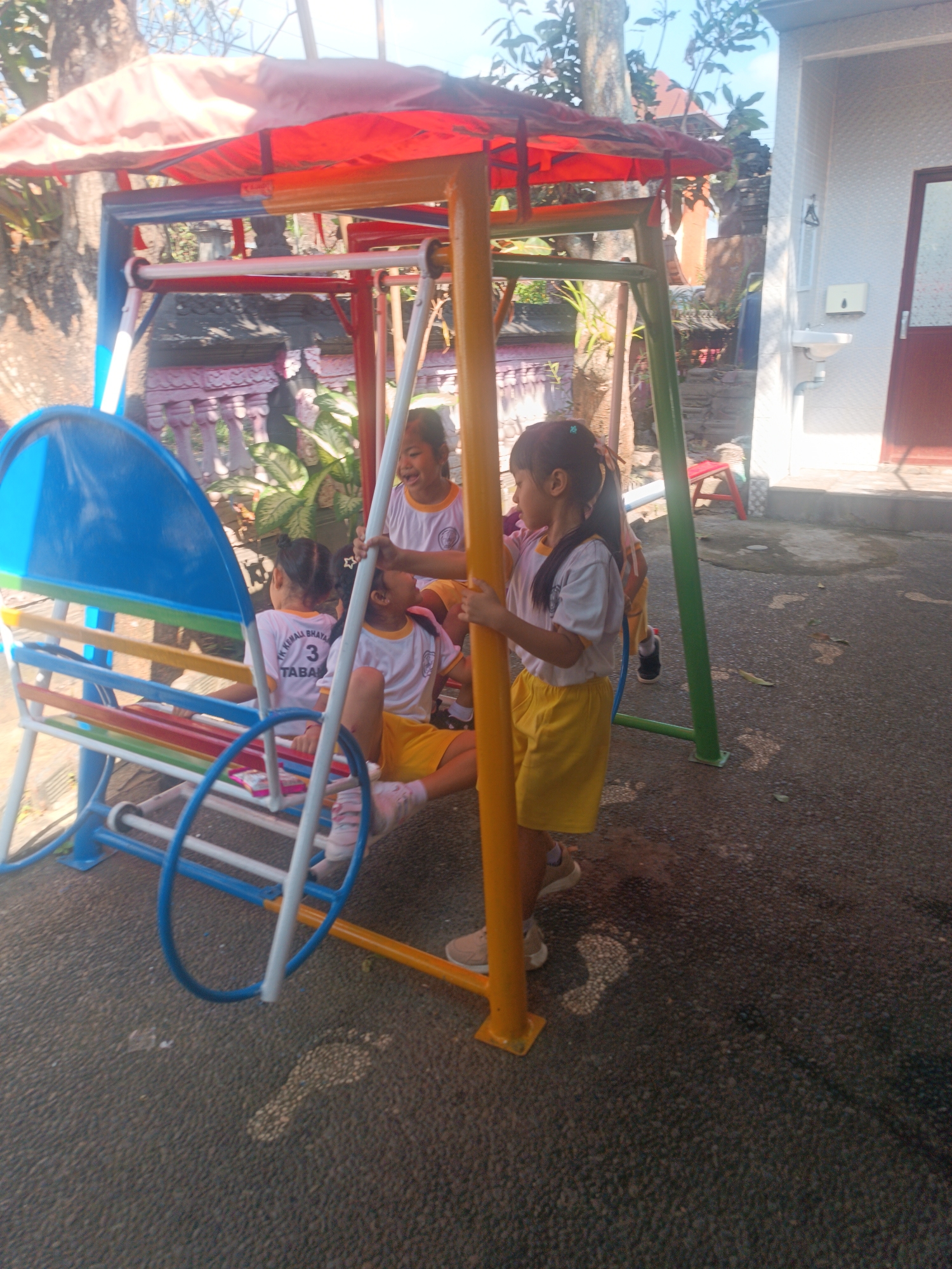 In the city of Tabanan, Indonesia, four young girls are engaged in play at an outdoor playground. The playground structure is composed of colorful metal poles and a red canopy. Three girls are seated on a multi-colored swing set, while a fourth girl stands next to them, holding onto a yellow pole. All four girls are wearing matching white shirts with a logo and yellow shorts. The ground surface is a textured asphalt with scattered leaves and a footprint marking. Behind the playground, there is a building with a white tiled exterior and a red door. The scene is illuminated by natural sunlight, casting shadows on the ground.