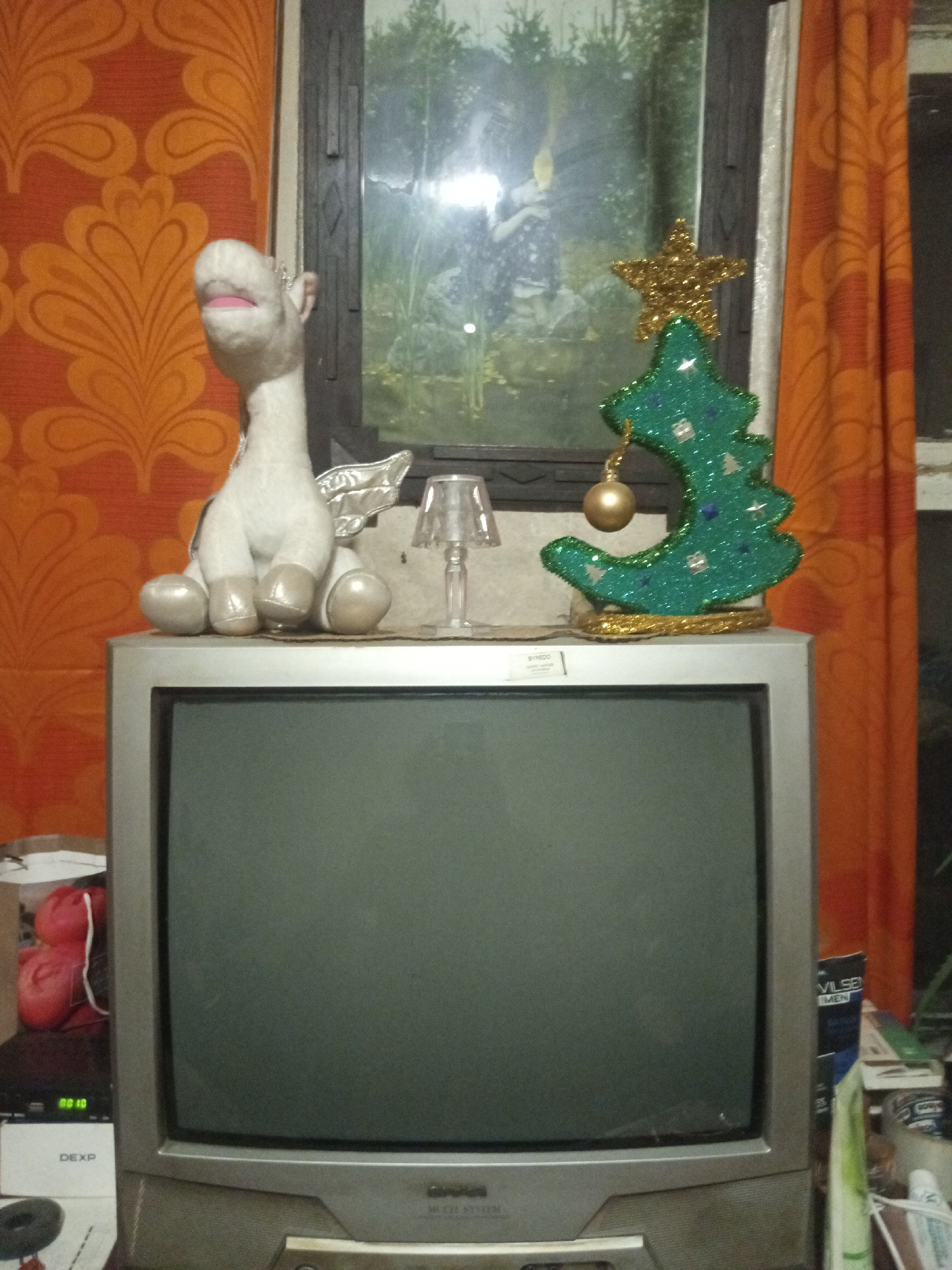 The image shows a vintage television set topped with various decorations. On the left side of the television, a plush unicorn toy with silver wings is seated. To the right of the unicorn, a small, clear lamp with a decorative shade is present. Next to the lamp, a glittery green Christmas tree ornament with a gold star on top and a gold ball hanging from it sits on the television. The background features a patterned orange wallpaper to the left and orange curtains to the right, with a window visible in the center showing trees and sky. The television itself is a CRT model with a dark grey screen. Additional objects are visible on a surface to the left of the television, including what appears to be a red plush toy and some electronic equipment with a display showing "00:18". The scene appears to be indoors, likely in a residential setting.