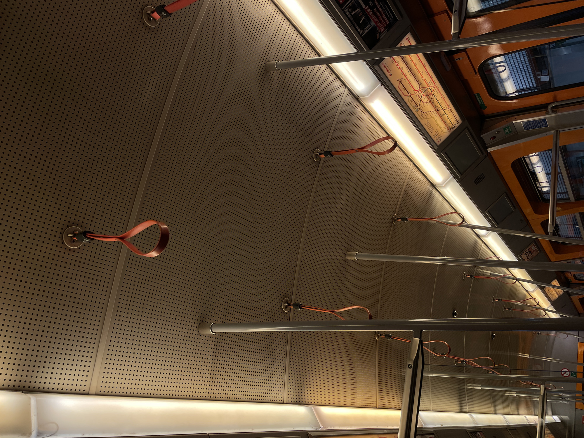 Here is a forensic description of the image:
**Setting:** The interior of a public transportation vehicle, possibly a subway car.
**Architecture and design:** The interior features a perforated metal ceiling with support bars and hand straps. The ceiling panels appear to be tan or beige in color. There are lights embedded within the ceiling structure. Along the upper right edge of the image, there is an advertisement, part of an orange and black window frame, and information signage.
**Objects:**
* Support Bars: Several metal bars run horizontally across the image.
* Hand Straps: Orange or reddish-brown loops are attached to the ceiling, likely for passengers to hold onto while standing.
* Perforated Metal Ceiling: The ceiling is made of metal with small circular perforations.
* Advertisements/Signage: There's a framed advertisement along the top edge.
* Lights: Integrated lighting fixtures run along the length of the ceiling.
**Light and shadow:** The scene is illuminated with artificial light, creating highlights on the ceiling panels and casting shadows from the support bars and straps.
**Location:** The signage mentions "Wiener Linien," suggesting the image was taken in Vienna (Wien), Austria.