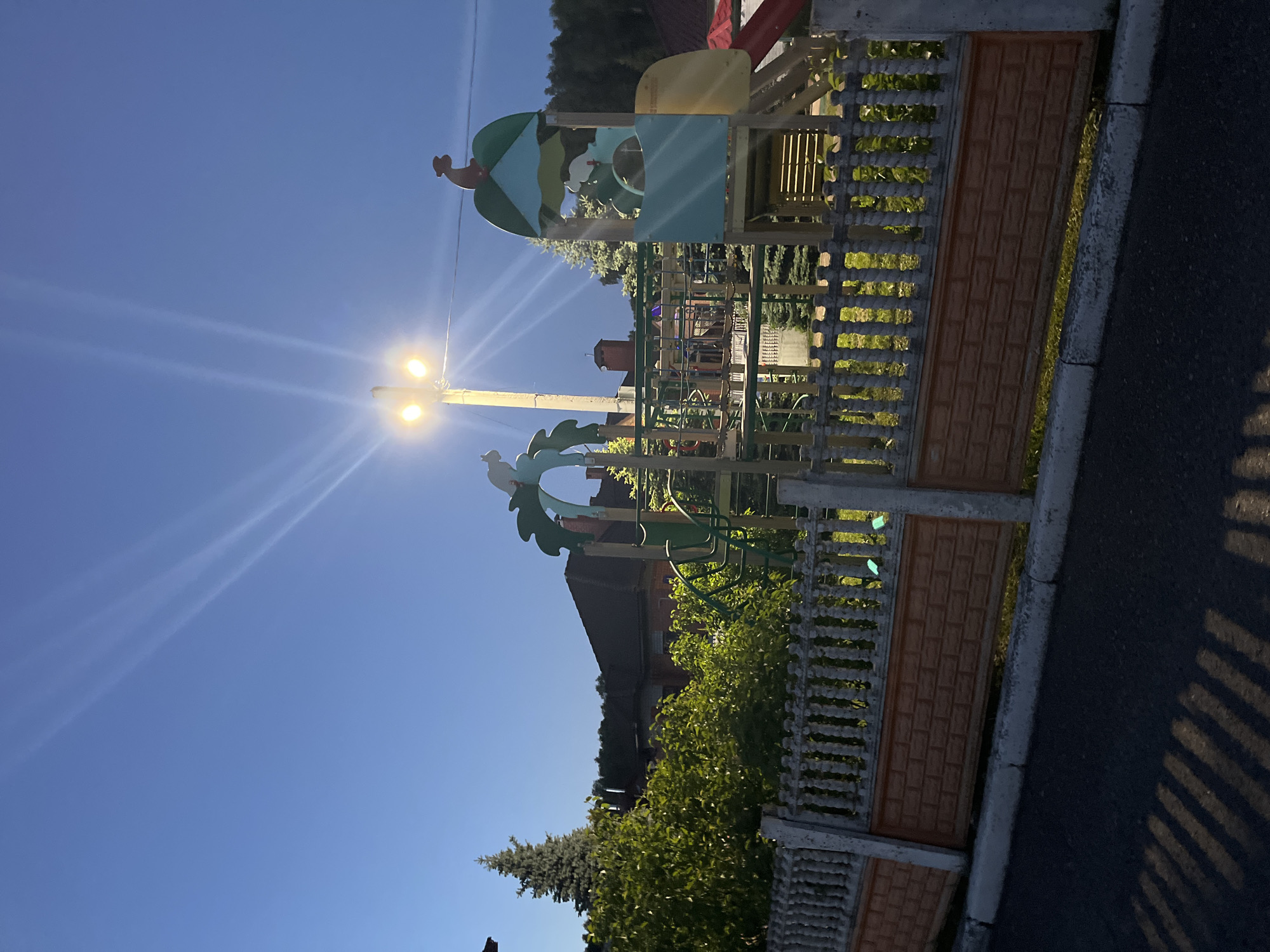 The image presents a playground adjacent to an asphalt road, likely situated in Ореховка, Russia. The scene is captured during either dusk or dawn, as evidenced by the bright blue sky complemented by an illuminated street lamp casting distinct light rays and prominent lens flares. Long, parallel shadows from a decorative fence stretch across the asphalt road.
The playground features a multi-level structure with colorful green, yellow, and blue elements, including slides, platforms, and climbing components. Sculpted motifs resembling dragon-like or bird-like figures adorn the top and lower sections of the playground equipment. A robust fence, composed of an ornate concrete base and textured, brick-patterned panels, separates the playground from the road.
In the background, lush green foliage from bushes and trees is visible, along with glimpses of residential buildings. The road has a clear concrete curb. The sky is largely clear and blue, with faint, wispy clouds barely discernible in some areas. No people are present in the image, and no visible text can be identified. The artificial glow of the street lamp adds a striking contrast to the natural ambient light.