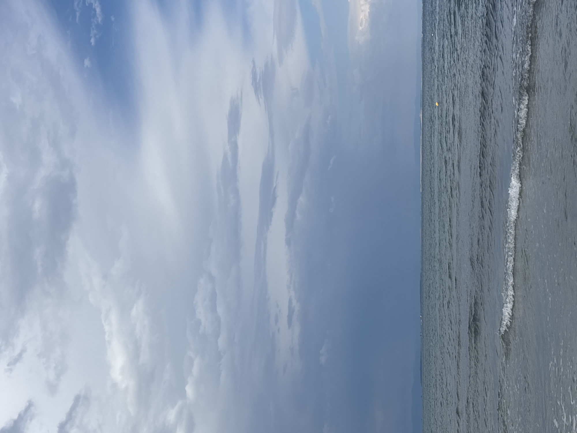 The image shows a view of the sea with a cloudy sky, taken from the coast of Mandra, Greece. The water is a deep blue-grey, with small waves lapping at the shore. The sky is filled with a variety of clouds, ranging from light and wispy to dark and heavy, suggesting overcast conditions. In the distance, a faint line of land is visible on the horizon, possibly mountains or a coastline. There is a small yellow buoy floating in the water. The overall scene is peaceful and serene, with a sense of natural beauty.