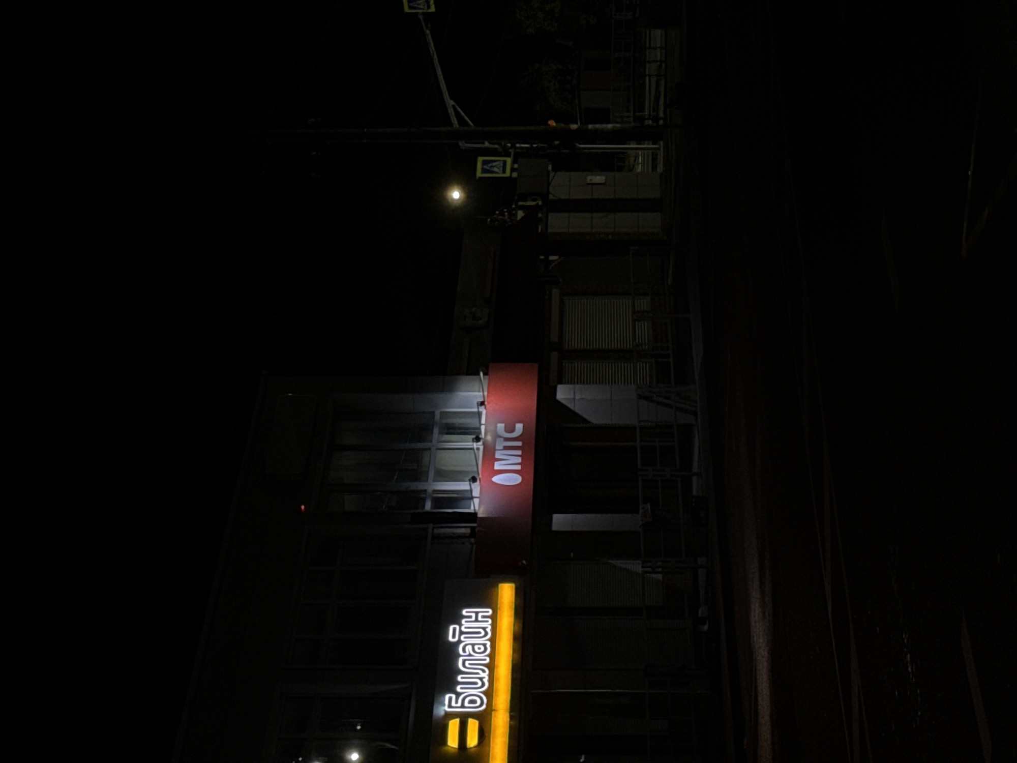 The image depicts a street scene at night in the city of Abinsk, Russia. On the left side of the frame, a building facade features two illuminated signs. The sign on the left is yellow with a black symbol resembling a hamburger bun and the Cyrillic text "Билайн" (Beeline), a telecommunications company. To its right, a red sign displays a white oval logo with a white Cyrillic text "MTC" (Mobile TeleSystems), another telecommunications company. The building appears to have large windows with dark shutters or blinds. In the background, a streetlamp with a bright full moon visible in the sky is present. There are also pedestrian crossing signs visible on a traffic light pole. The street in front of the building is dark, with some faint reflections of light on the wet asphalt. There are no people visible in the image.