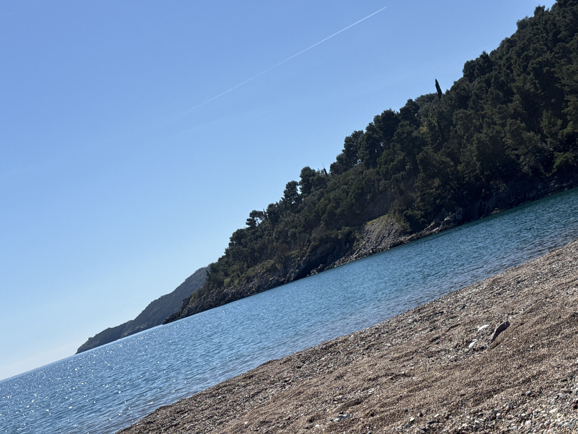 The image captures a coastal scene with a beach composed of pebbles leading up to a body of water. A forested hill or landmass rises from the water's edge, extending upwards and covering the right side of the image. The sky is clear and blue, with a contrail visible from an aircraft. The location is in the city of Unknown, Unknown.