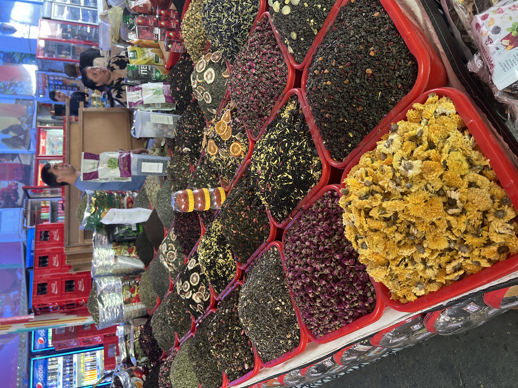 A market stall in Tashkent, Uzbekistan, displays a vibrant selection of dried herbs, flowers, and teas. The stall is laden with large red plastic bins filled with various loose-leaf products. In the foreground, a bin is brimming with dried yellow chrysanthemum flowers, and next to it, a bin contains dried rosebuds. Further back, more bins hold a variety of teas and herbs, some mixed with dried fruit pieces like orange slices. Several plastic bags and boxes of packaged tea are also visible on the stall. In the background, a man in a blue shirt and a woman in a patterned shirt are interacting at the stall. Behind them, refrigerators are stocked with drinks, and pallets of Coca-Cola crates are stacked. The overall scene depicts a bustling marketplace with an abundant display of goods.