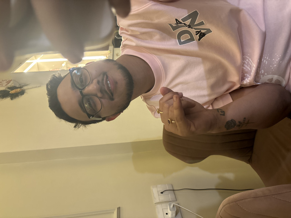 This image captures a young adult male in an indoor, well-lit setting, likely a cafe or private residence in Howrah, India. The subject, of South Asian descent, has short dark hair, a light beard, and wears black-rimmed glasses. He is looking directly at the camera with a slight smile. He is dressed in a light pink short-sleeved t-shirt featuring a grey graphic with the letters "DEYN" and a stylized design on the chest.

His left arm is bent at the elbow, and his hand is partially raised, displaying two rings: a gold-colored ring with a dark red stone on the ring finger and a plain gold-colored band on the index finger. A tattoo is visible on his left forearm, depicting a black rose with faint text underneath and a "L+" or "L5" symbol above. A blurry white object, possibly a part of a phone or another device, is visible in the extreme upper right foreground, held by an unseen hand, suggesting the image is a selfie or a close-up taken by another person.

The background features a light beige wall. On the left, two white electrical outlets, consistent with Indian standards, are installed, with a white power adapter plugged into the lower outlet and a black cable extending downwards. Behind the man, a brown upholstered chair with vertical stitching is visible. A fragment of another light-colored, possibly pink, upholstered chair is present on the far left. The upper right background includes indistinct decorative elements and a subtle reflection of light.
