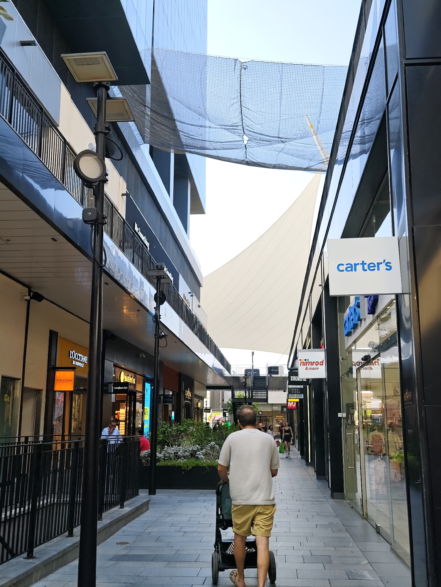 The image depicts a pedestrian walkway in Herzliya, Israel, likely within a modern commercial or shopping district. The walkway is paved with large, light-grey rectangular tiles laid in a staggered pattern. On the right side of the walkway, a building features large glass windows and a prominent white sign with blue text reading "carter's". To the left of the "carter's" sign, a smaller sign reads "nimrod & more". Further down the walkway on the right, there are storefronts with visible displays and mannequins.
On the left side of the walkway, a multi-story building with dark grey cladding and horizontal windows is visible. A black pole with attached lights and electrical boxes stands in the foreground. Above the walkway, a dark netting is suspended between buildings, and a white fabric canopy or sail is stretched across a portion of the sky.
In the foreground, a man is pushing a stroller along the walkway. He is wearing a light-colored t-shirt and khaki shorts. His back is to the camera. Further down the walkway, other individuals can be seen, including a person in a red shirt and another in a blue shirt in the background on the left. The lighting suggests it is daytime.