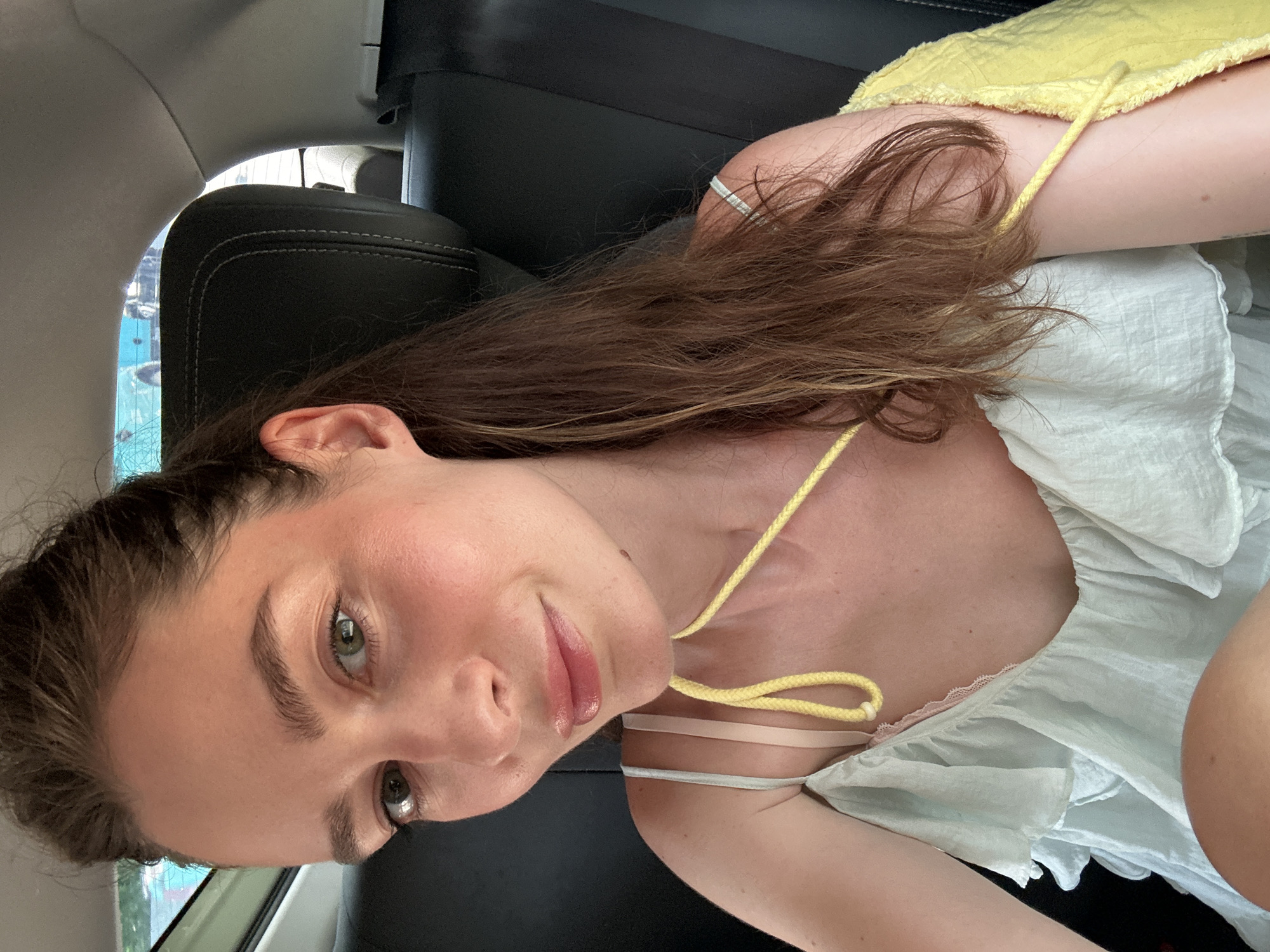 A young woman with fair skin, green eyes, and light brown hair is depicted in a photograph taken from a close-up, slightly angled perspective. The subject is likely seated in a vehicle, with a black leather car seat visible in the background. The woman wears a pale blue top with a yellow tie-strap detail and a hint of yellow fabric visible near her shoulder, suggesting a layered garment. The location in Choeng Thale, Thailand, is not directly discernible from the visible elements in the image, so it can only be specified if the vehicle window shows a view of the landscape.