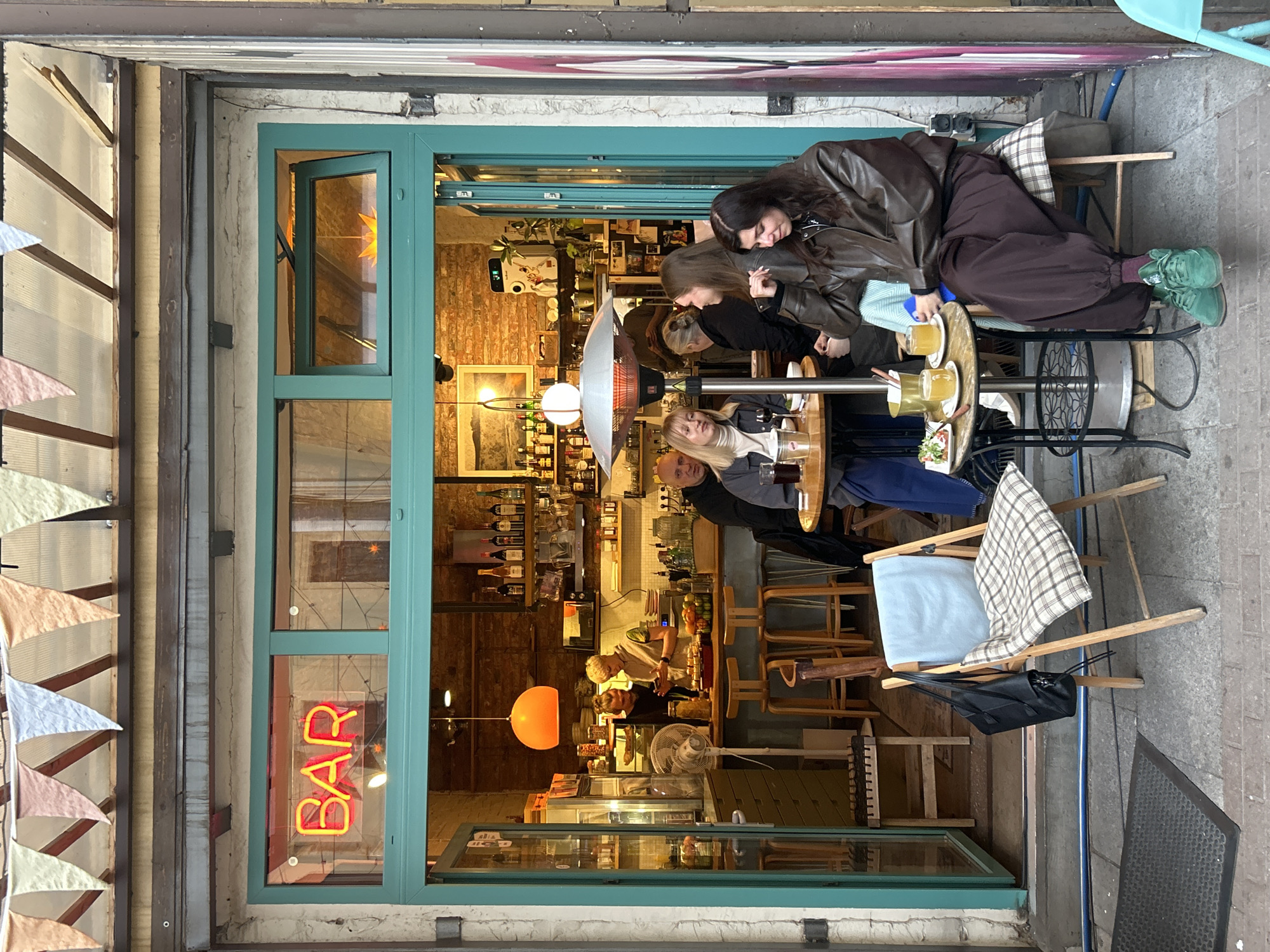 An outdoor bar in Saint Petersburg, Russia. A group of five people are seated at a small round table outside a bar, illuminated by a central heating lamp. The woman on the left has blonde hair and is wearing a black coat and blue jeans, the person next to her wears a black jacket. The woman in the middle has her hair in a bun, and she is wearing a black top. To her right, the other two women are wearing brown jackets. They are seated around the table, drinking. The bar's interior features shelves stocked with bottles and two employees, presumably bartenders, working behind the counter. A neon "BAR" sign is visible in the window. Pennant banners are attached to the side of the building.