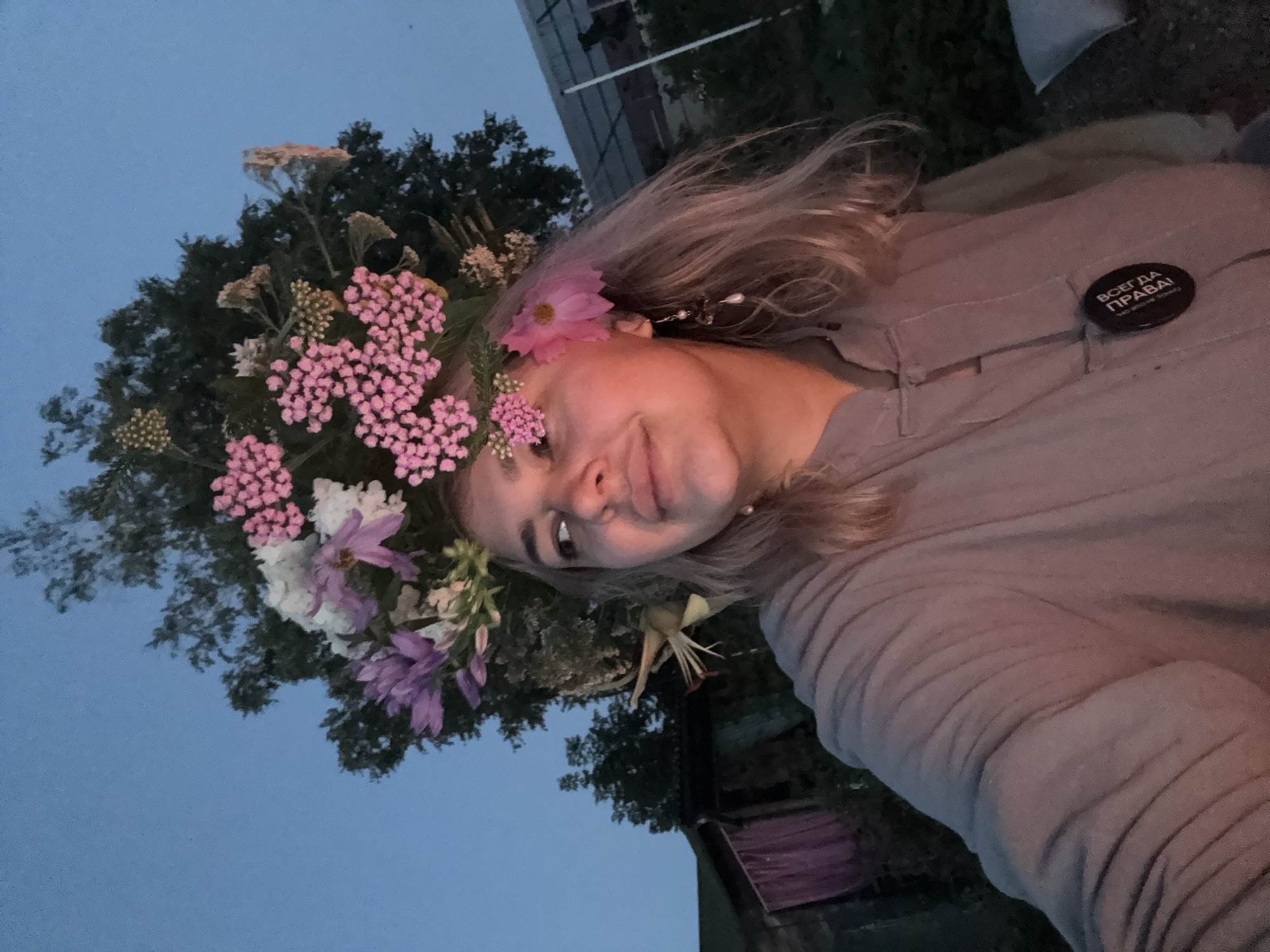 In an outdoor setting, a young woman with blonde hair and a floral headdress is photographed from the chest up, looking to her left. The headdress is large and elaborate, composed of various flowers and greenery, including pink clusters and purple and white blooms. She is wearing a muted, long-sleeved garment, possibly a tunic or a jacket, with a button closure. A black, circular pin with white text is attached to the garment, partially visible. She is also wearing an earring with beads and a pearl. The background suggests a rural or semi-rural environment with trees, a building structure, and what appears to be a fence or netting. The lighting indicates it is either dusk or dawn, with the sky a deep blue. There are no other individuals clearly visible in the frame, and the woman is looking slightly away from the camera, with a subtle smile. The location is identified as Birobidzhan, Russia.