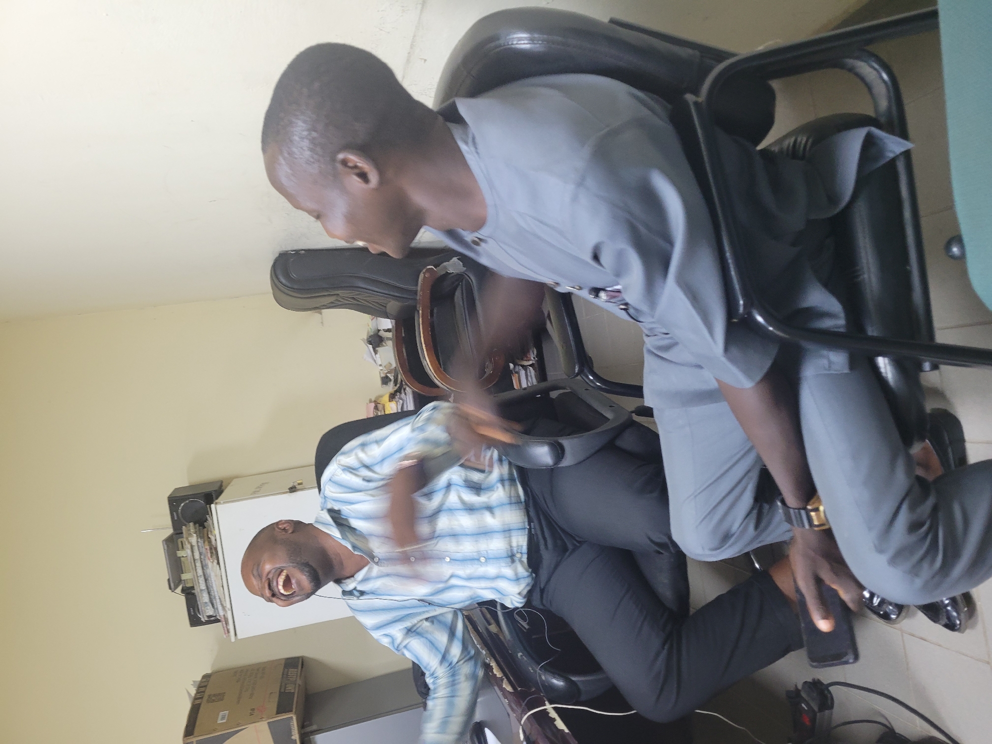 Two men are seated in an office environment, appearing to be engaged in a humorous conversation. The man on the left, wearing a blue and white striped shirt and black trousers, is laughing heartily with his head thrown back. He is seated in a black office chair with wooden armrests. To his right, the second man, dressed in a grey traditional outfit and grey trousers, is also looking to his left and appears to be smiling or laughing. He is seated in a similar black office chair. The room has cream-colored walls and tiled flooring. Various office items are visible, including a filing cabinet with a radio on top, a cardboard box, and what appears to be a computer desk with a keyboard and monitor on the left. The overall atmosphere suggests a casual and lighthearted moment within a typical office setting in Abuja, Nigeria.