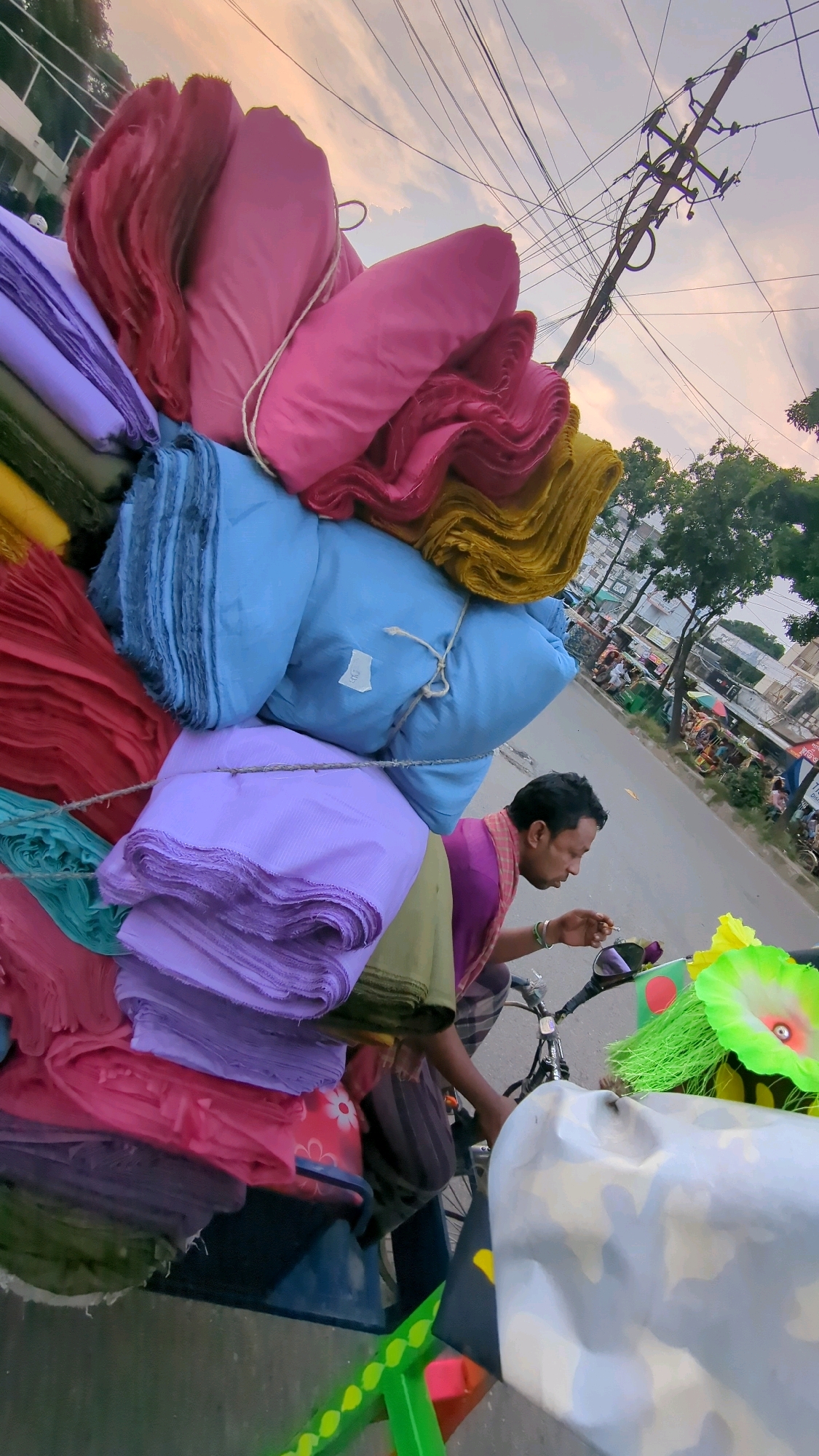 In Dhaka, Bangladesh, a man rides a bicycle laden with a large stack of colorful textiles. The fabrics, neatly folded and piled high, include shades of pink, purple, blue, red, and yellow. The man, wearing a traditional lungi and a scarf draped around his neck, appears to be smoking a cigarette as he navigates the street. The bicycle also carries a large, camouflaged object, possibly a seat cover or a large bag, in the foreground. In the background, a typical urban scene unfolds with buildings, trees, other vehicles, and overhead power lines suggesting a busy street in the city. The sky is overcast, with the soft light of either dawn or dusk filtering through the clouds.