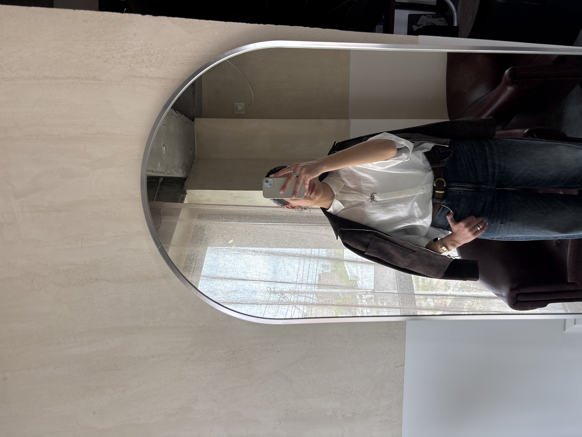 The image displays a mirror selfie taken indoors, reflecting a person, part of a room, and an exterior view, set in Yerevan, Armenia. A large, rounded rectangular mirror with a silver frame is the central element.
The subject in the reflection is a person holding a light-colored smartphone, possibly an iPhone, covering most of their face except for their lips. They are wearing a white collared shirt, dark blue jeans, and a dark brown jacket draped over their shoulders. A dark brown belt with a prominent gold-toned buckle is visible at their waist. Accessories include multiple silver-colored rings on their fingers, a gold-toned watch with a square face on their left wrist, and a silver-colored necklace with a stylized "C" or crescent-shaped pendant. A small, embroidered logo, potentially a horse and rider design, is subtly visible on the left chest of the white shirt.
The interior reflected in the mirror shows light-colored walls, some with a visible texture resembling concrete. A white electrical outlet with a plug is on one wall, alongside what appear to be steps or architectural ledges. To the right of the person, a dark brown, possibly leather, armchair or seat is reflected. Beyond the interior, a large window covered by sheer, light-colored curtains reveals an outdoor urban landscape with distant buildings and some greenery, indicating it is daytime.
The activity captured is a casual mirror selfie. Notable visible text is limited to the stylized "C" on the necklace and a small, unidentifiable logo on the shirt. The overall setting appears modern and well-lit by natural light.