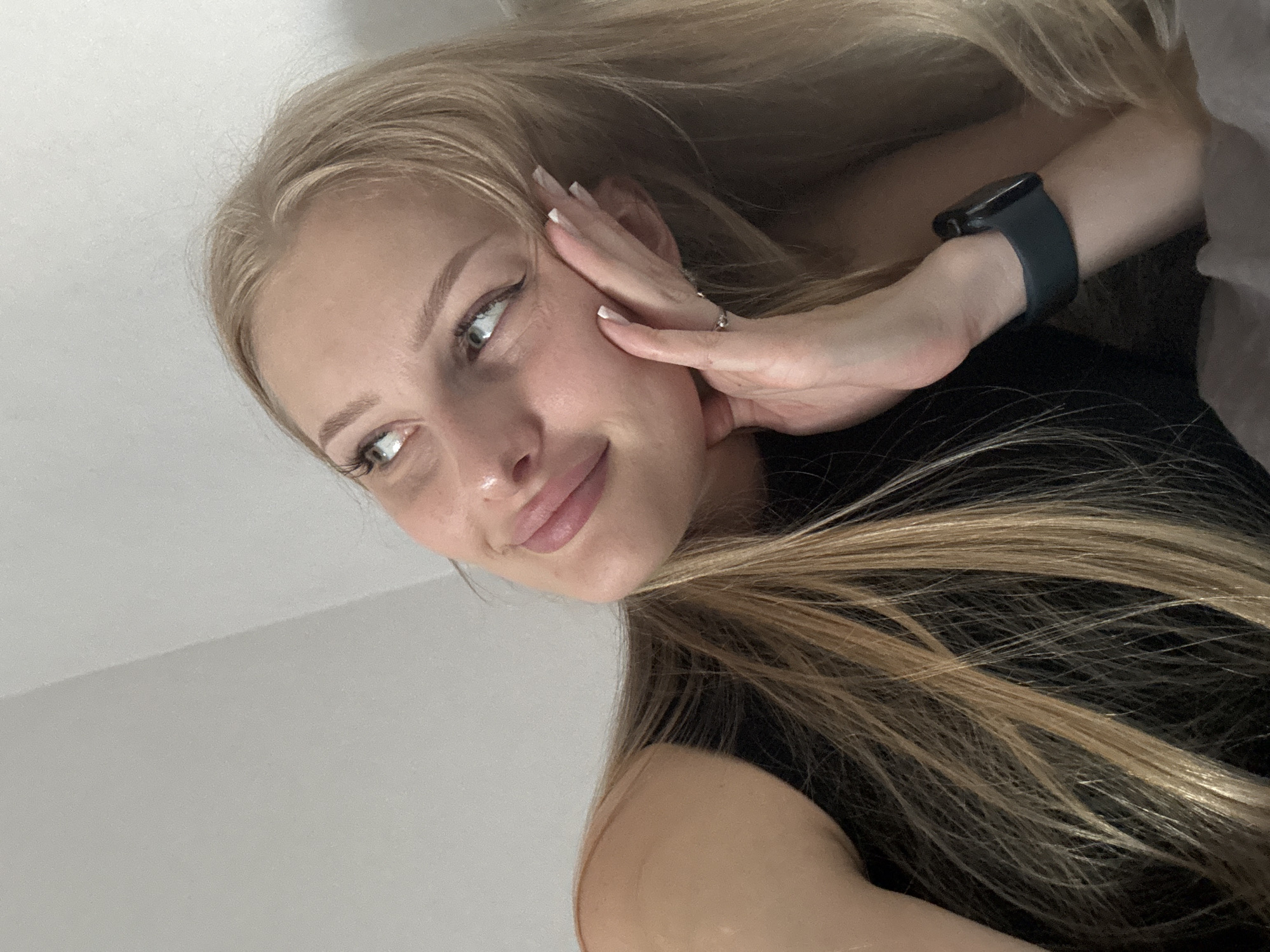 A young woman with blonde hair and light blue eyes is captured in a close-up, head-and-shoulders shot. She is looking upwards and to her right, with her right hand resting on her cheek and chin. She is wearing a black sleeveless top and a dark smartwatch on her left wrist. Her nails are manicured with white tips. The background consists of a plain, light-colored wall with a subtle angled shadow. The lighting appears to be soft and diffused. The woman has a thoughtful or wistful expression. There is no indication of the city of Perm, Russia, or any specific urban environment in the image.