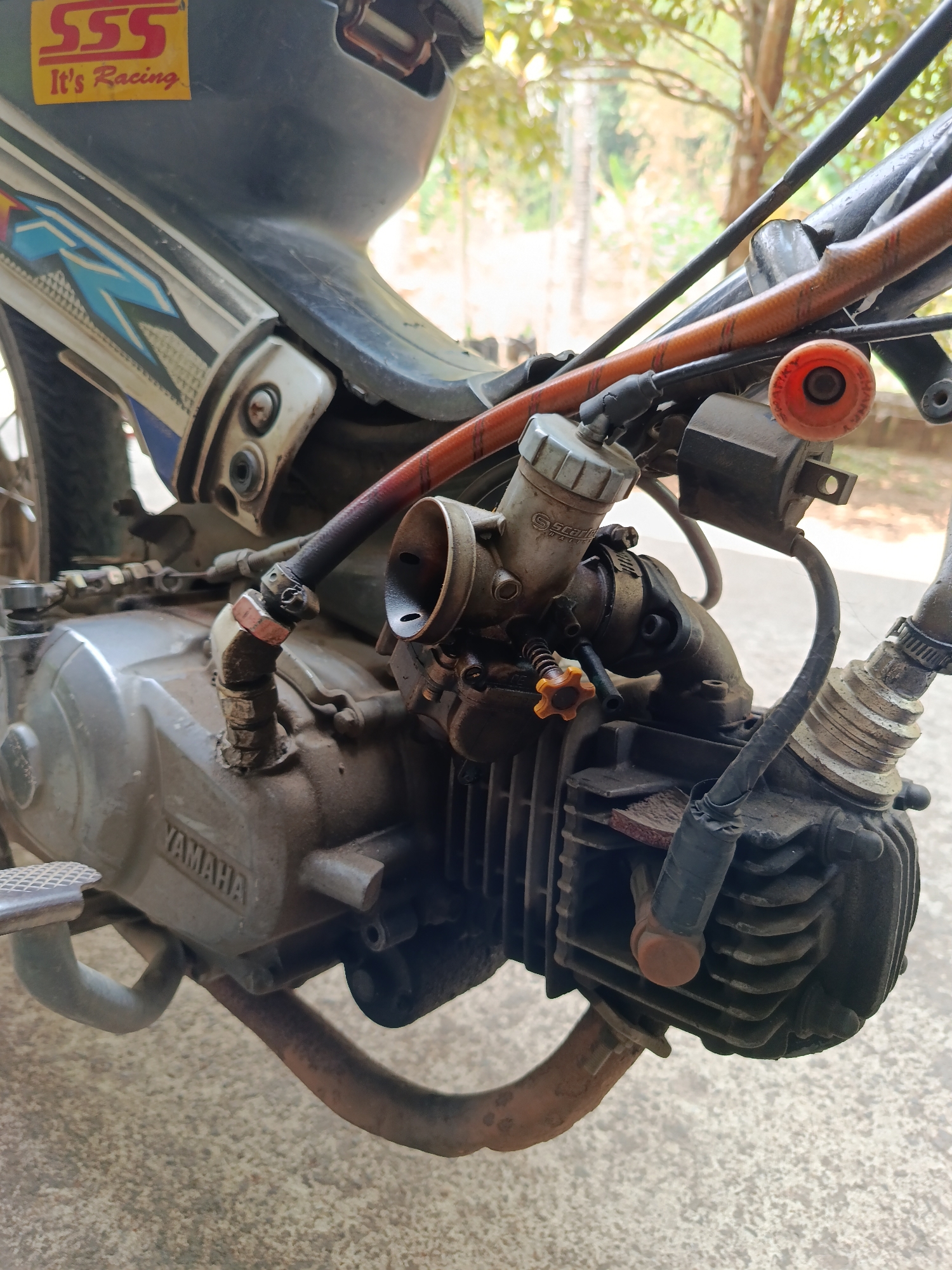 The image shows a close-up view of a motorcycle engine, with the brand name "YAMAHA" visible on the engine casing. The engine appears to be older and somewhat dirty, with visible wear and tear on some components, including rust on the exhaust pipe. A part of the motorcycle's bodywork, featuring a "SSS It's Racing" sticker, is also visible in the upper left corner. The background is blurred, but suggests an outdoor environment with trees and foliage. The scene is likely a roadside or a yard in Lempong, Indonesia, given the natural lighting and vegetation. There are no people or animals present in the image.