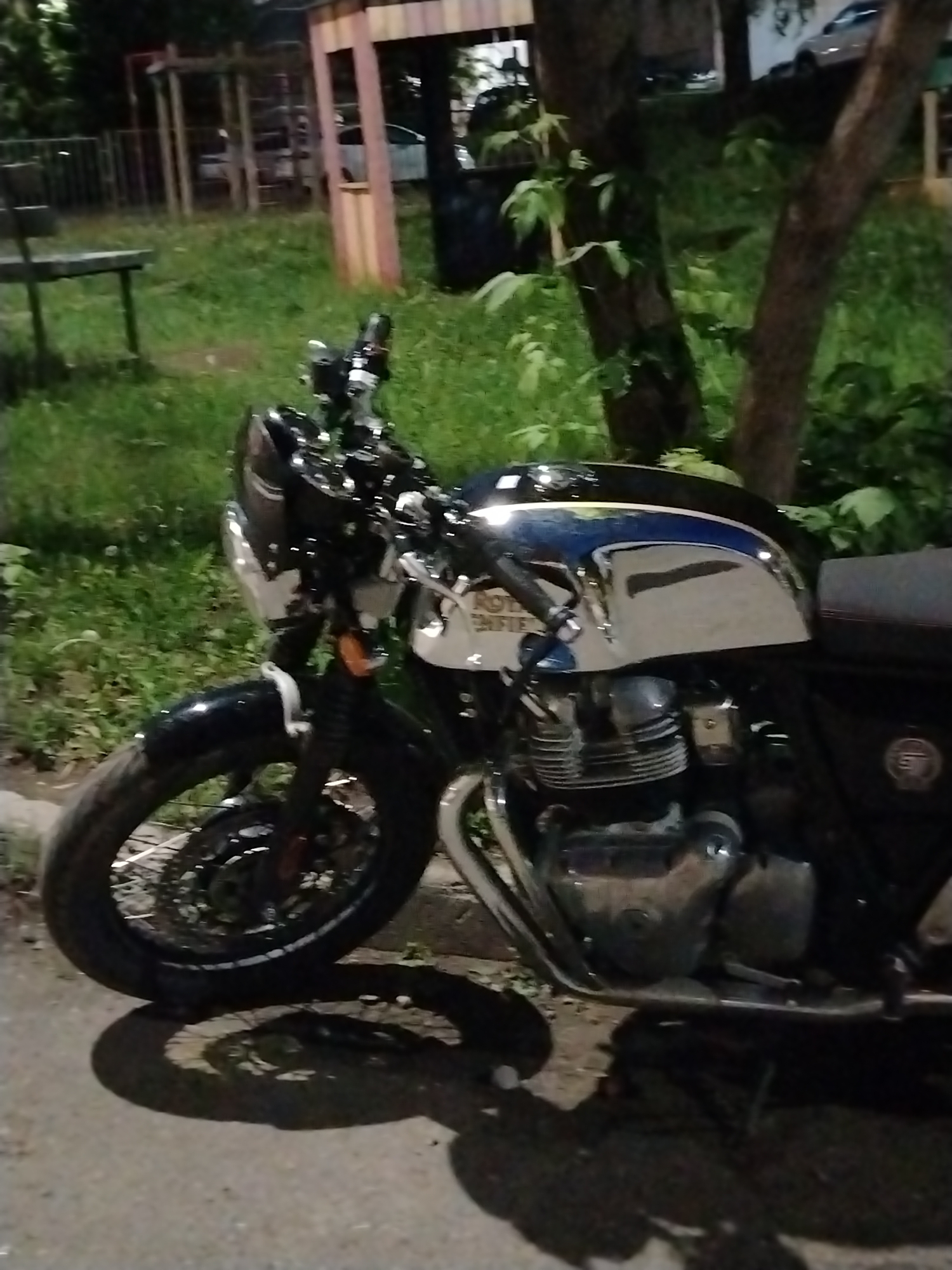 The image shows a motorcycle parked on a paved surface, likely a sidewalk or parking area. The motorcycle is a modern classic style, with a chrome fuel tank and black accents. It is positioned in front of a grassy area with trees and a wooden playground structure in the background. The location is possibly in Izhevsk, Russia. The time of day appears to be evening or night, given the lighting conditions. No people are visible in the image.