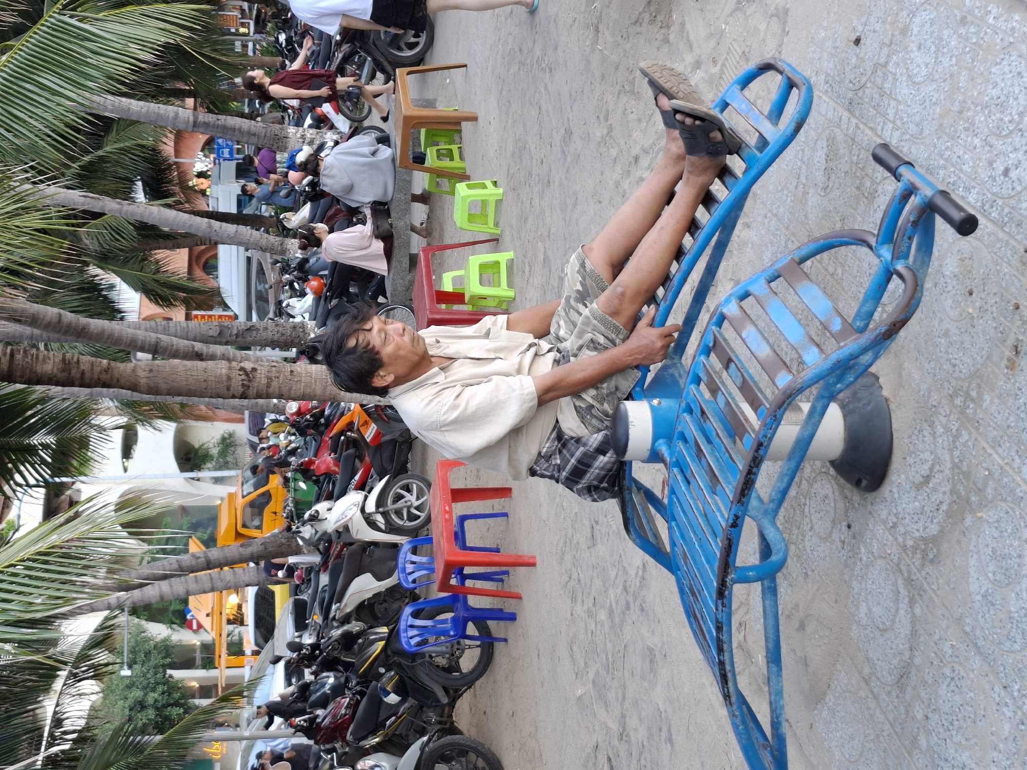 A man is sitting on a blue metal exercise apparatus on a paved area with sand. He is wearing a light-colored short-sleeved shirt and camouflage shorts. Palm trees are in the background, and several motorcycles are parked. In the distance, there is a yellow food truck and buildings. The scene appears to be a street or a public area in Bac Nha Trang Ward, Vietnam.