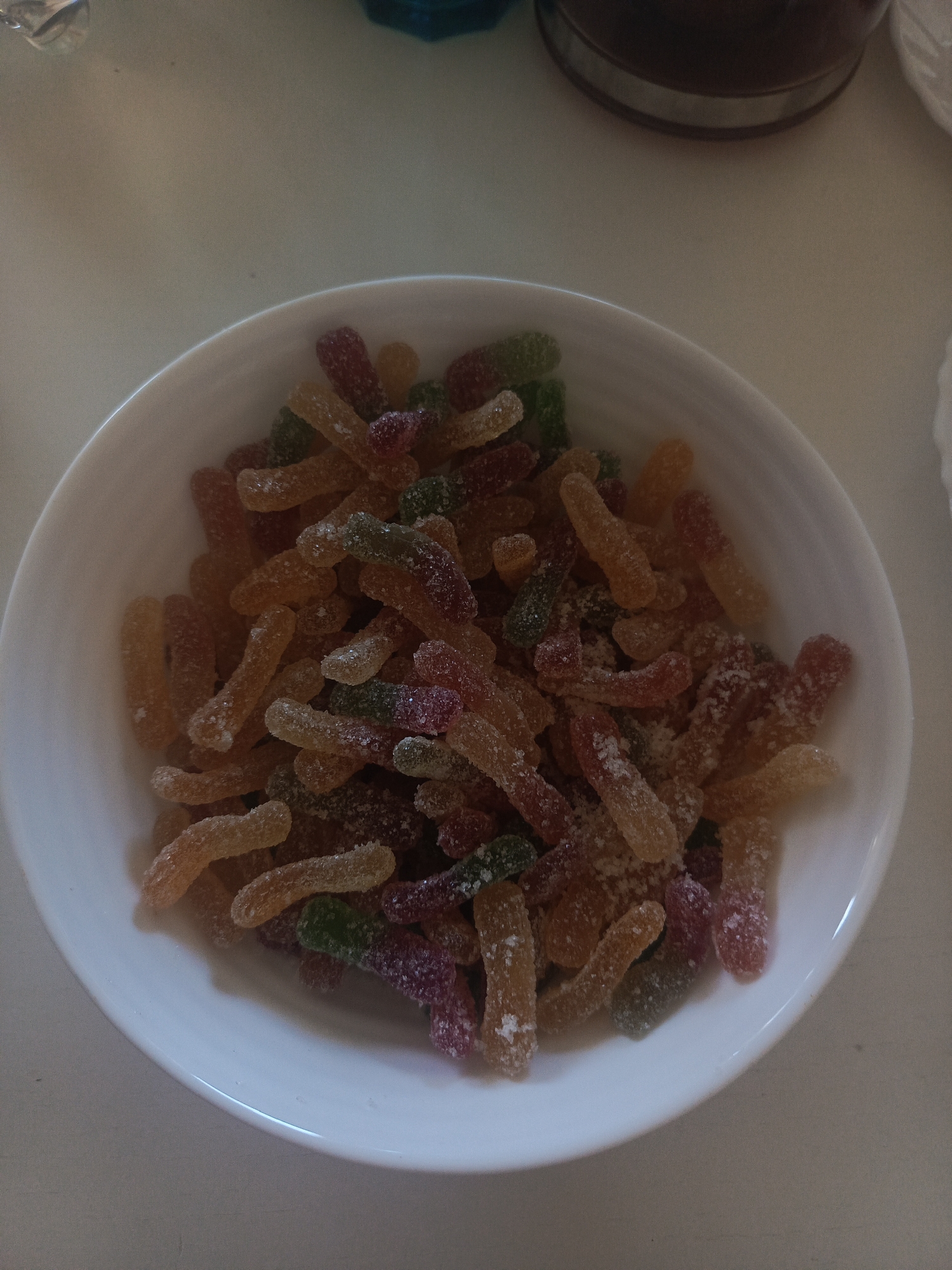 A white bowl is filled with a pile of colorful, worm-shaped candies coated in a sugary or crystalline substance. The candies appear to be a mixture of orange, red, green, and purple hues. The bowl sits on a pale, neutral surface. Parts of other items are visible at the edges of the frame, a jar and another unidentified object.