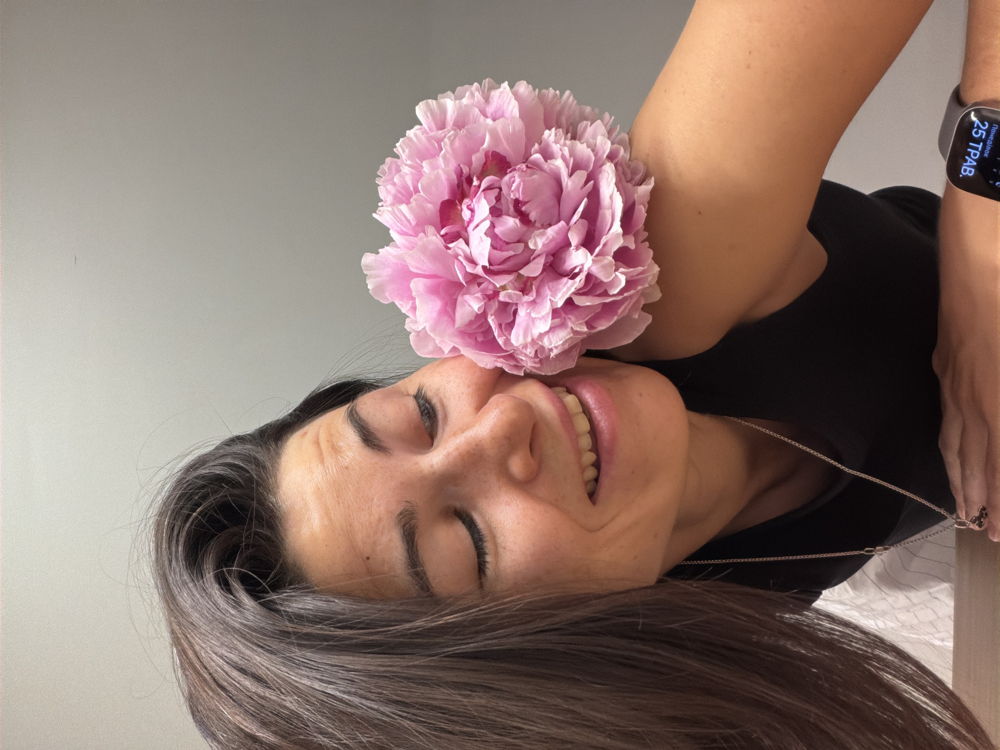 The image depicts a woman with long, dark hair, lying on her side and smiling with her eyes closed. A large, pink peony flower is positioned on her shoulder. She is wearing a black top and a silver necklace. A smartwatch is visible on her wrist, displaying the date "25 ТРАВ." The background is a plain, light gray wall. There is no indication of the location being Budva, Montenegro.