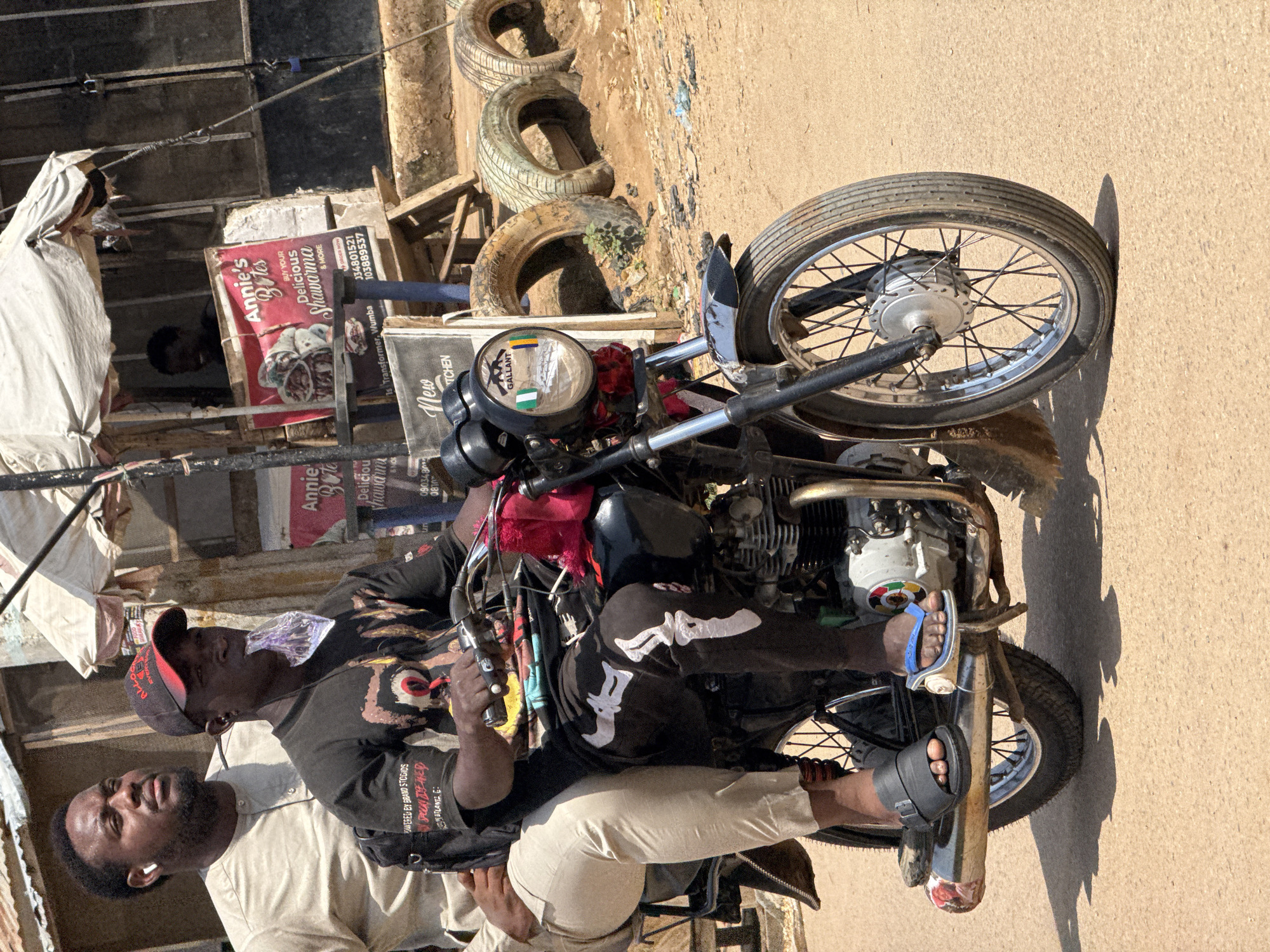 Two men are riding on a motorcycle in Abuja, Nigeria. The man in front is wearing a black t-shirt and a red cap and is holding a plastic bag in his mouth. The man behind him is wearing a light-colored, long-sleeved shirt and has earbuds in his ears. They are on a dirt road, and in the background, there is a food stall with a sign that says "Annie's Bites." There are also several tires stacked behind the stall. The scene appears to be a busy street market or roadside vendor area.