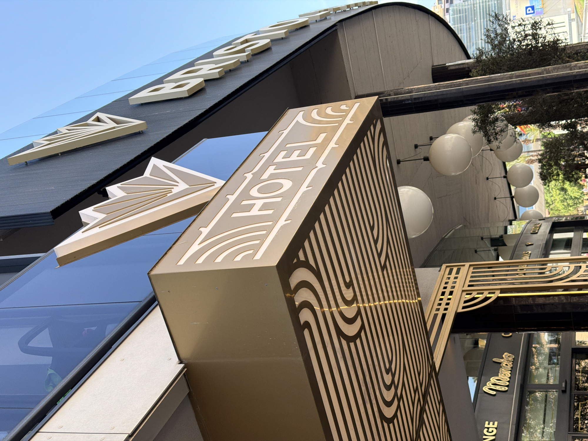The image shows the exterior of a hotel in Benidorm, Spain. The hotel has a modern architectural design with a curved facade and large glass windows. A prominent golden sign displays the word "HOTEL" in a stylized font. Above the sign, a decorative element features geometric lines resembling a fan. On the building's facade, large golden letters spell out "BRISTOL," indicating the hotel's name. Below the "HOTEL" sign, a patterned canopy with black and gold stripes is visible. Further down, another storefront sign reads "Moncho" in a cursive gold font. Several large white spherical light fixtures are suspended from the ceiling of the hotel's entrance area. The sky is clear and blue. Trees and some buildings are visible in the background.