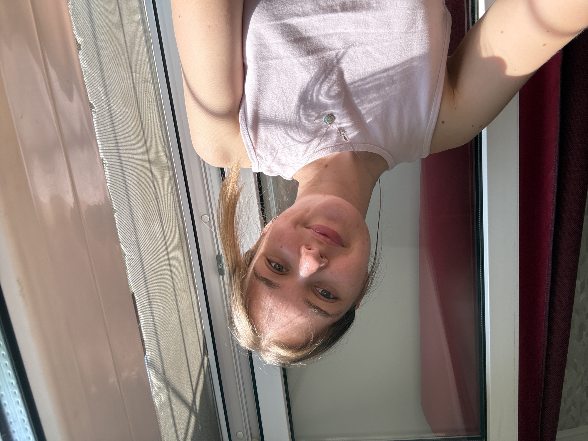 A young woman with blonde hair and wearing a pink tank top is posing for a selfie. She is standing in front of a window, likely indoors. The window has a screen and a curtain. Red curtains are partially visible on the right side of the image. Based on the visible clues, it is not possible to confirm the specific location in the city of Perm, Russia.