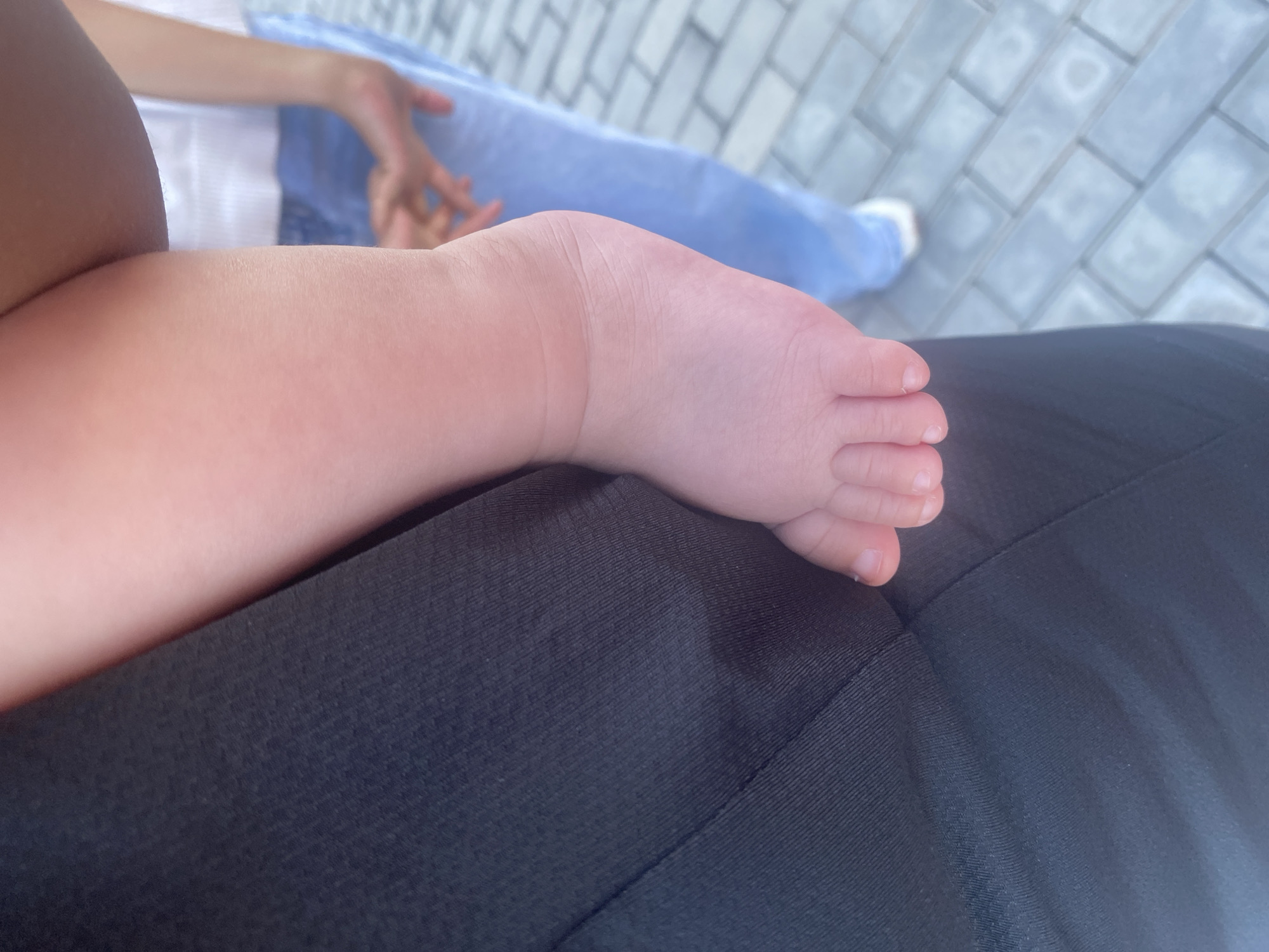 A close-up, waist-level shot shows the bare foot of a baby resting against the dark fabric of an adult's clothing. The baby's skin is pink and soft, with clearly defined toes and fingernails. The adult's clothing appears to be made of a textured material, possibly athletic wear. In the background, out of focus, another person is visible wearing a white top and blue jeans, standing on a paved surface. The paving stones suggest an outdoor urban setting, potentially in Manas, Kyrgyzstan, though this location is not explicitly confirmed by visual cues within the image.