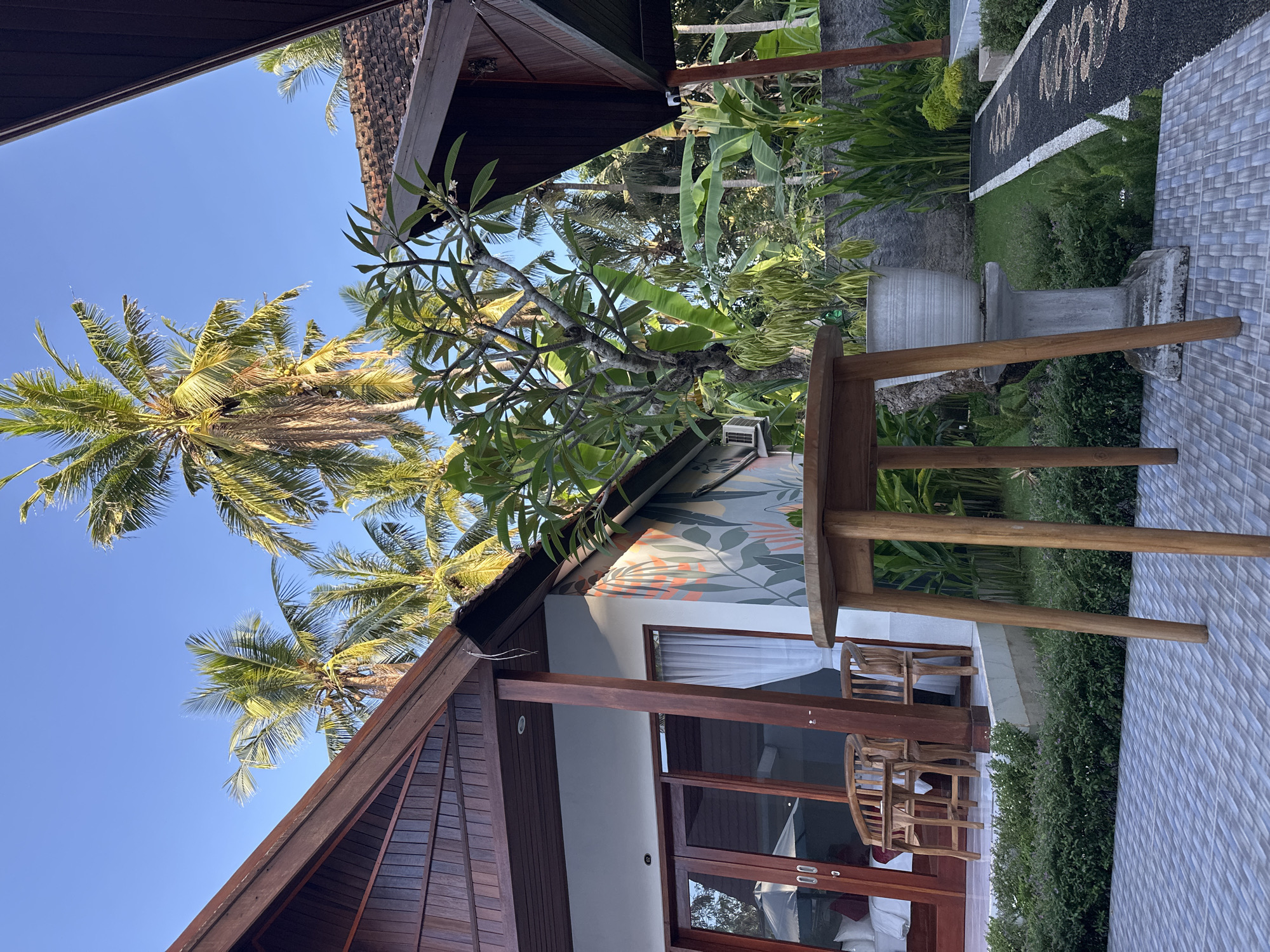 The image shows a view of a building with a wood-paneled roof in Nusa Penida, Indonesia. In the foreground, there is a wooden table on a tiled patio. The building has glass doors and windows, with a mural painted on the wall above. Palm trees are visible in the background against a clear blue sky. Lush greenery surrounds the building.