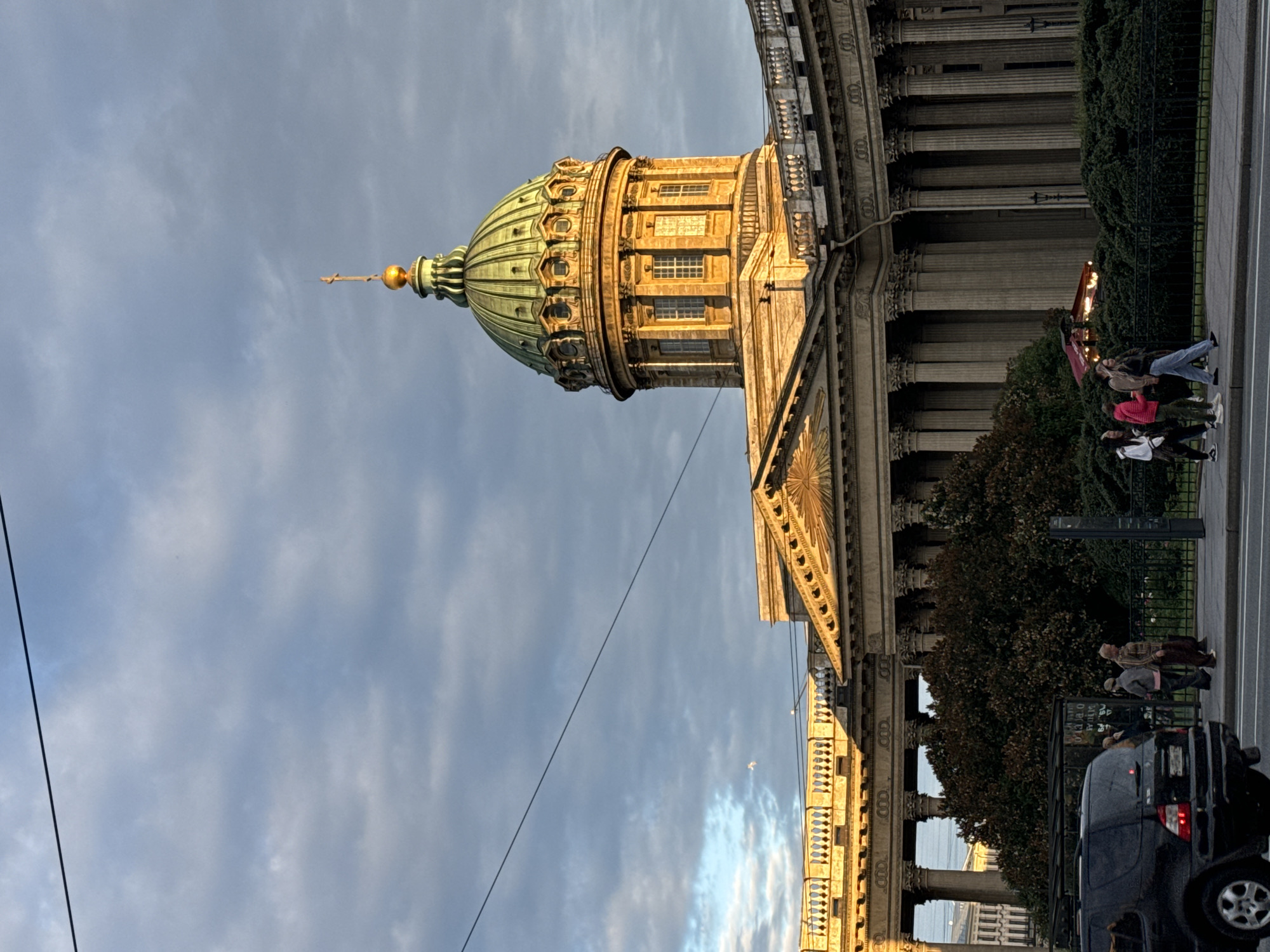 The image shows the Kazan Cathedral, a prominent landmark in Saint Petersburg, Russia. The cathedral's large green dome, topped with a golden cross, dominates the upper half of the frame. The facade of the cathedral features a colonnade of classical columns, bathed in the warm, golden light of the setting sun. In the foreground, a black SUV is parked on the left side of the street. Several people are walking on the sidewalk in front of the cathedral, some near a bus stop shelter. The sky is partly cloudy, with streaks of blue sky visible between the clouds.
