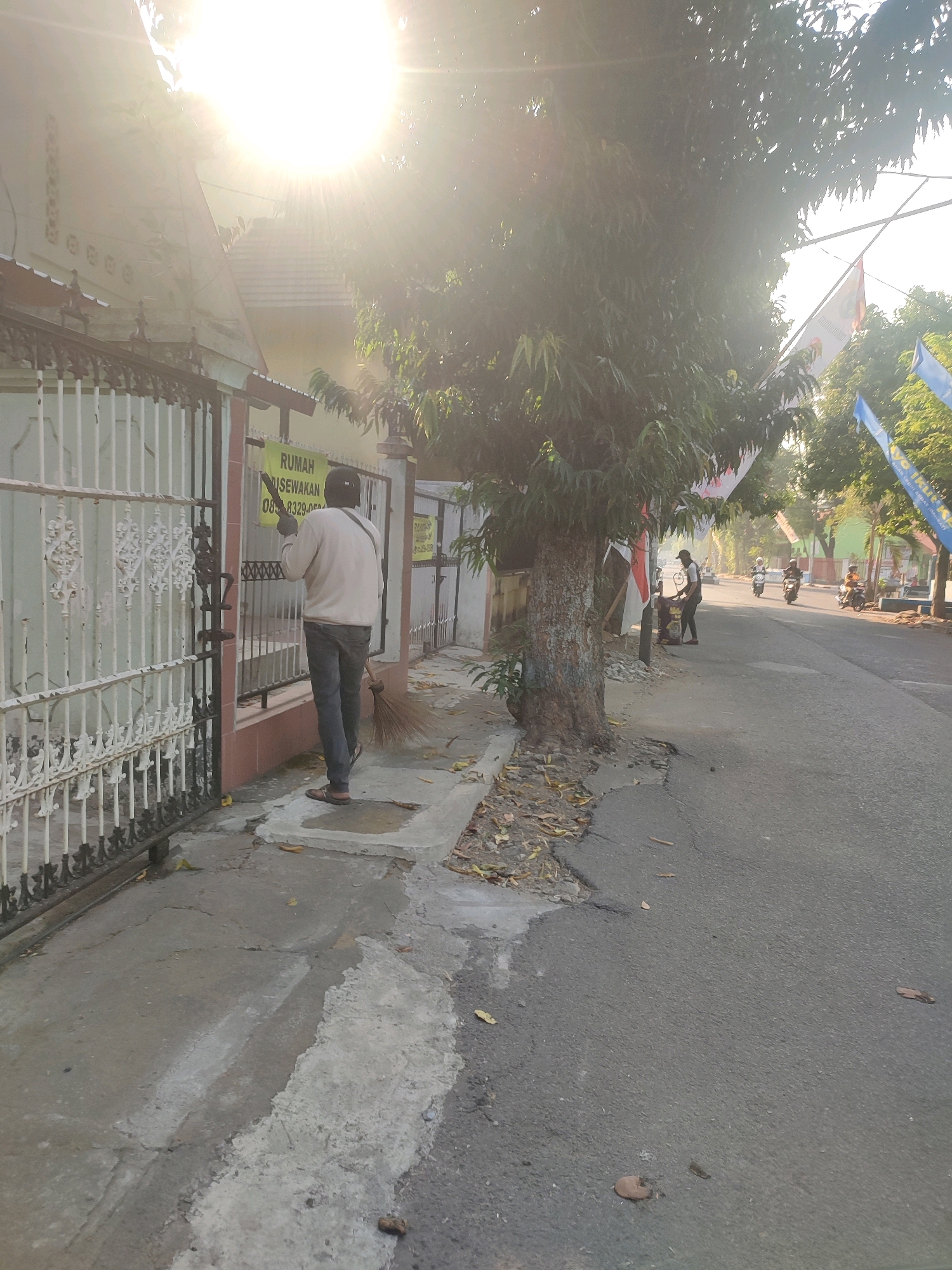 The image depicts a street scene in Nganjuk, Indonesia, likely in the morning given the strong sunlight and lens flare from the lower left. A person is standing on a sidewalk in front of a gated property, sweeping the ground with a broom. The person is wearing a light-colored long-sleeved shirt, dark pants, a cap, and sandals. A yellow sign is attached to the gate, indicating a house for rent. To the right of the sidewalk, a road runs parallel to it, with a few motorcycles and riders visible in the distance. Trees line the street, and flags are hung from poles along the side of the road. The environment appears to be a residential or mixed-use area.